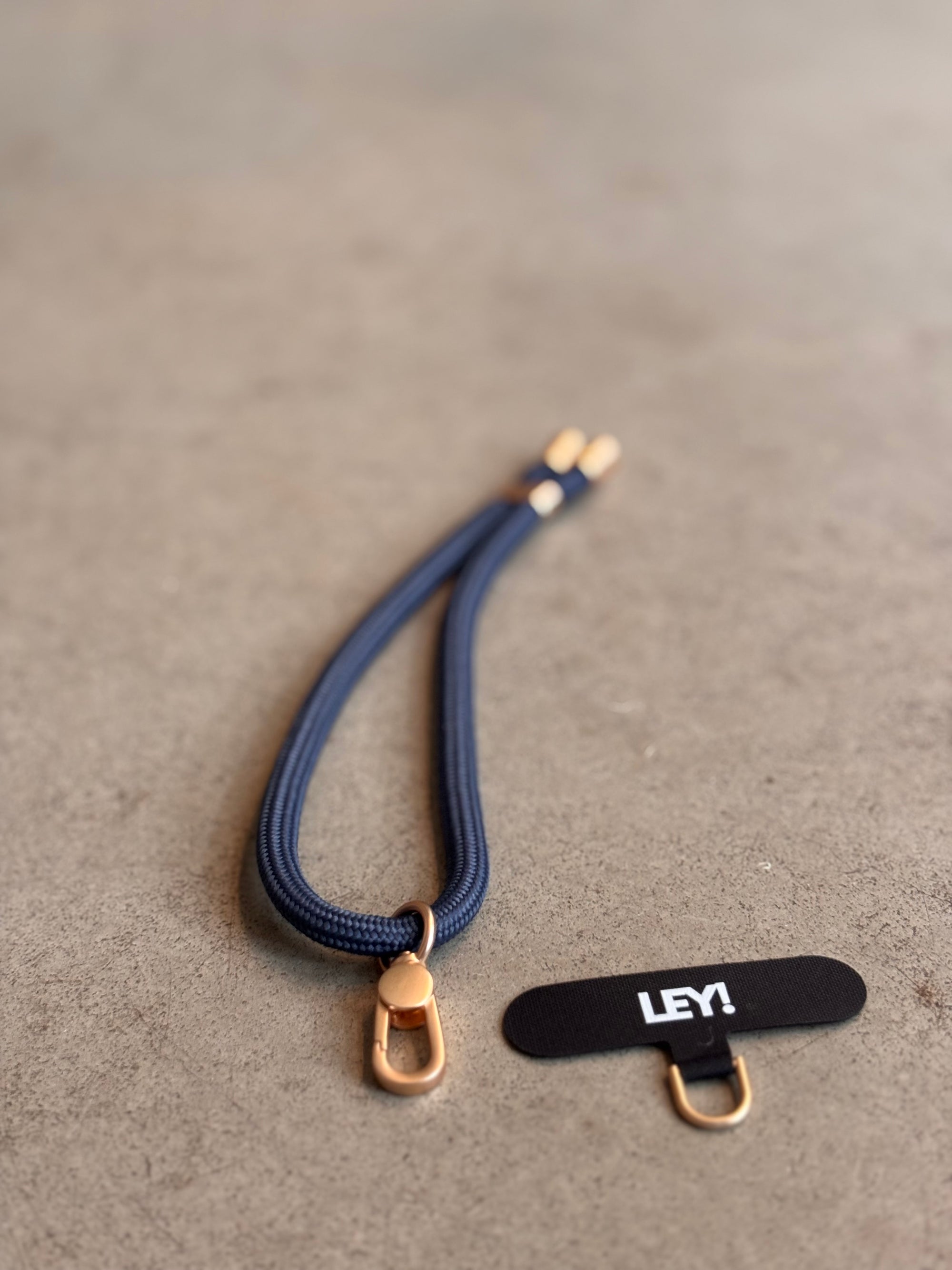 Ein navyblauer LEY! Schlüsselanhänger mit goldener Hardware liegt auf grauem Untergrund neben einem schwarzen Etikett mit weißer "LEY!"-Aufschrift und sorgt dafür, dass dein Handy immer griffbereit ist.