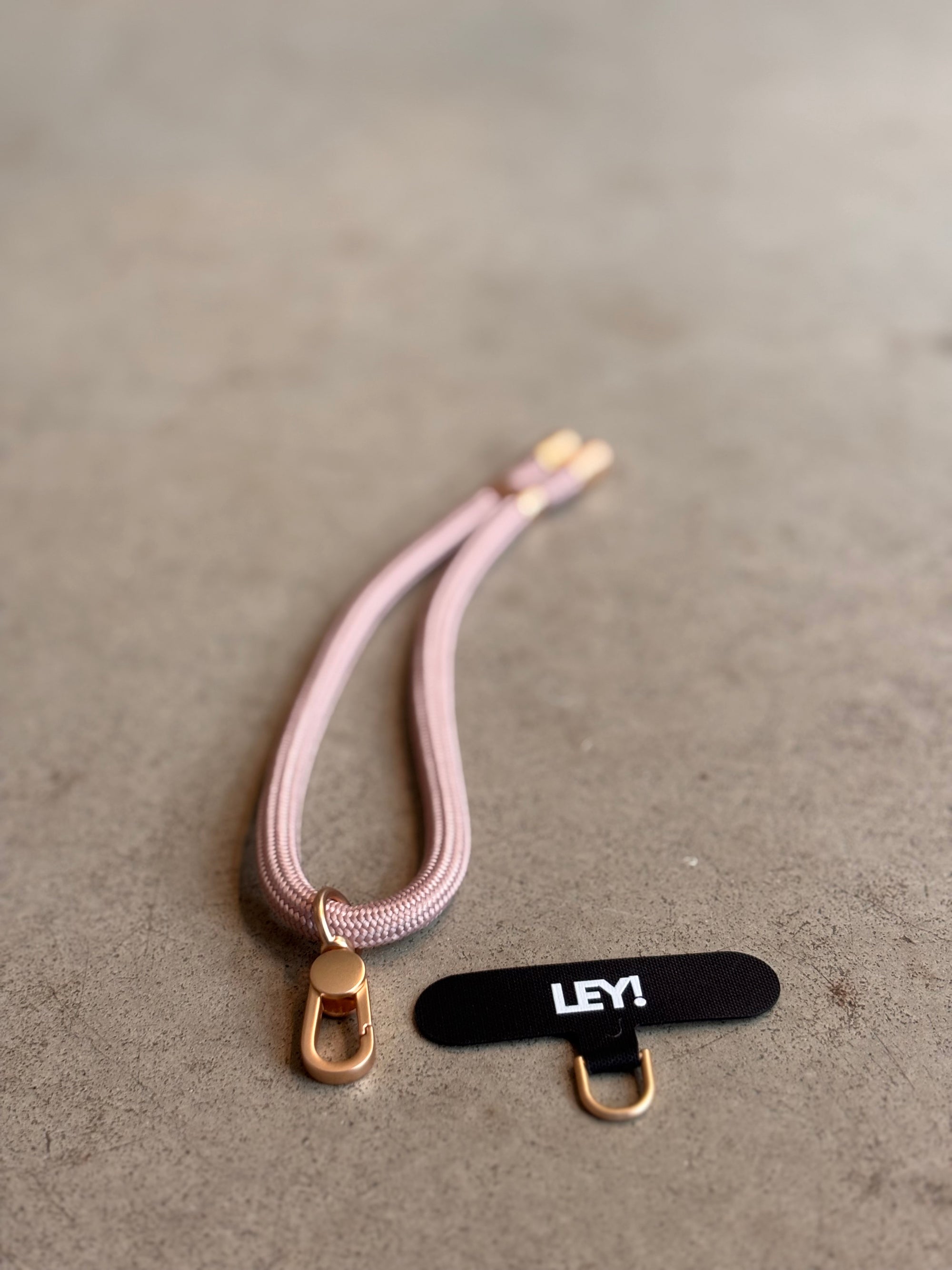 Ein rosa-goldener LEY! Schlüsselanhänger mit Metallspangen liegt auf einer grauen Fläche neben einem kleinen schwarzen "LEY!"-Anhänger mit einem goldenen Ring - ideal zum schnellen Finden und Tragen deiner Schlüssel in trendigen Farben.