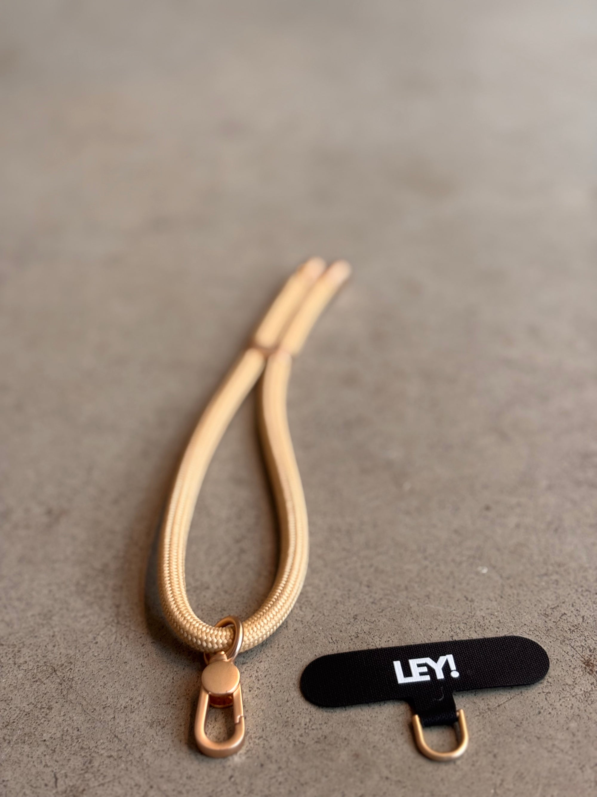 Ein goldener Schlüsselanhänger von LEY! mit einer klobigen Schließe ruht auf einer grauen Oberfläche neben einem schwarzen Etikett - ein unverzichtbares Accessoire, um dein Handy in trendigen Farben stilvoll zur Hand zu haben.