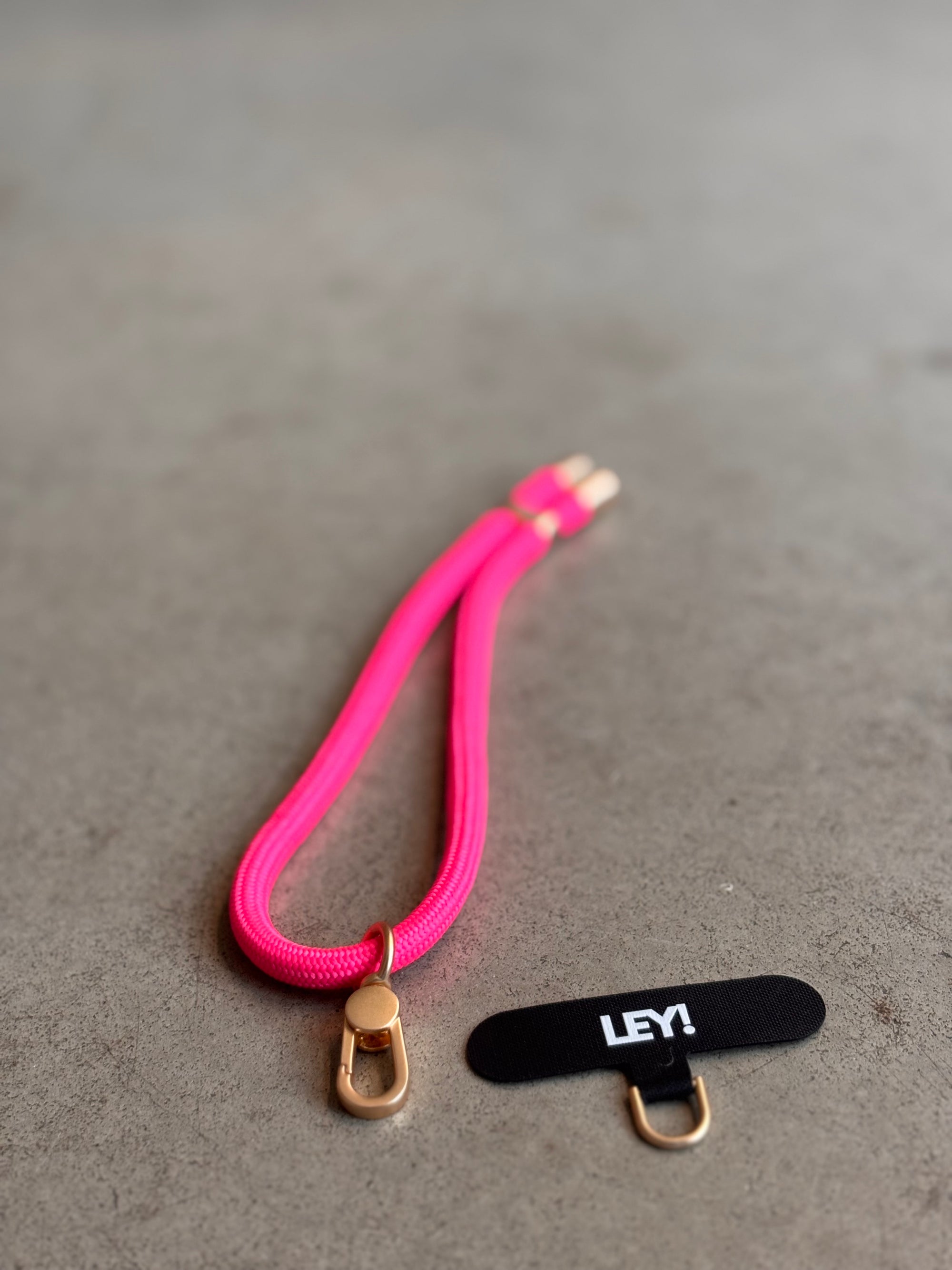 Ein leuchtend pinker Schlüsselanhänger von LEY! mit goldenen Verschlüssen ruht auf einer grauen Fläche neben einem schwarzen LEY!-Stoffetikett und einem goldenen Ring - so bleibt dein Handy stets griffbereit.