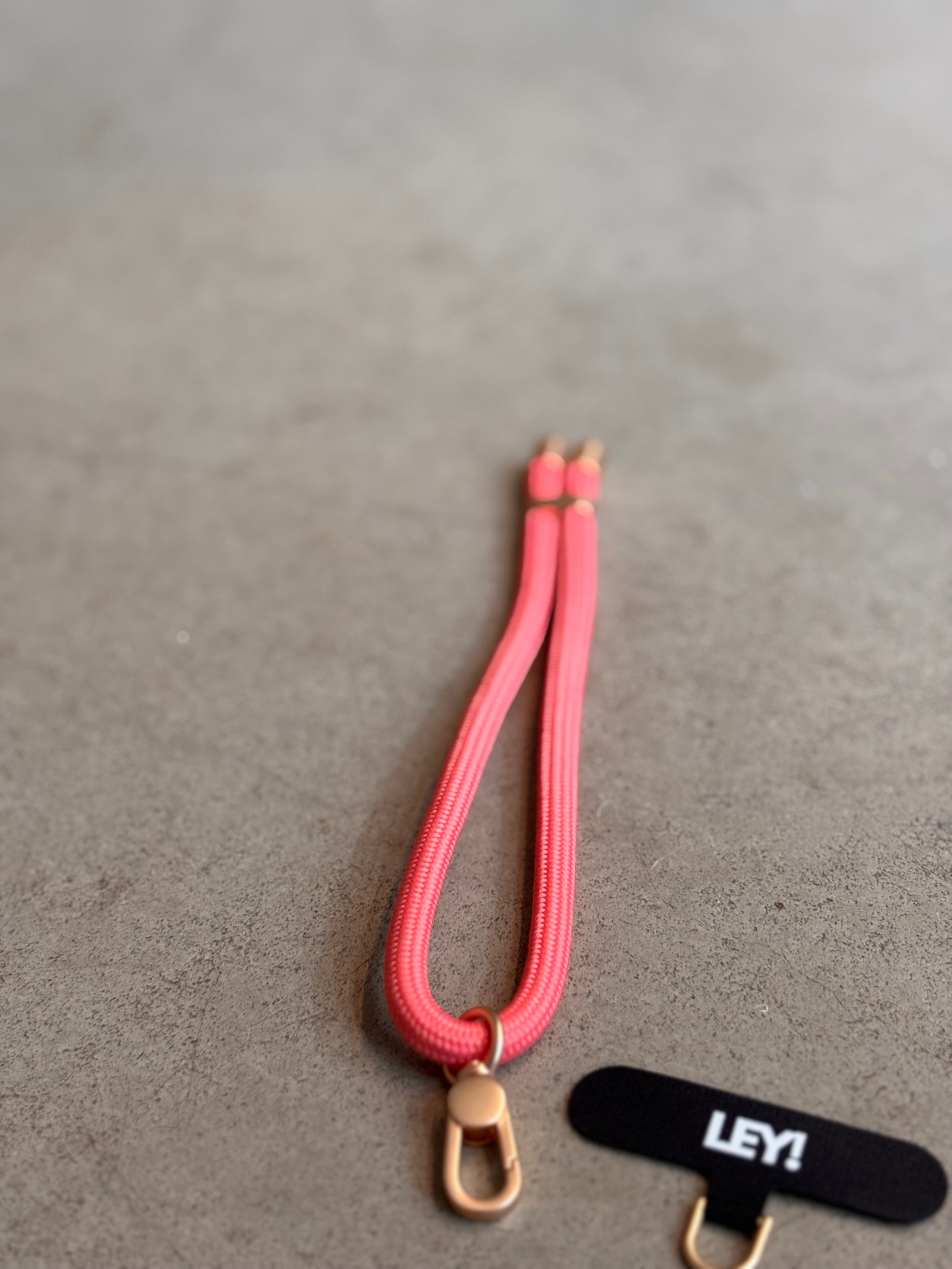 Ein rosafarbener Schlüsselanhänger von LEY! mit goldenen Beschlägen ruht auf einer grauen Fläche neben einem schwarzen Stoffanhänger mit "LEY!" in Weiß-Praktisch, um dein Handy immer griffbereit und in trendigen Farben bei dir zu tragen.