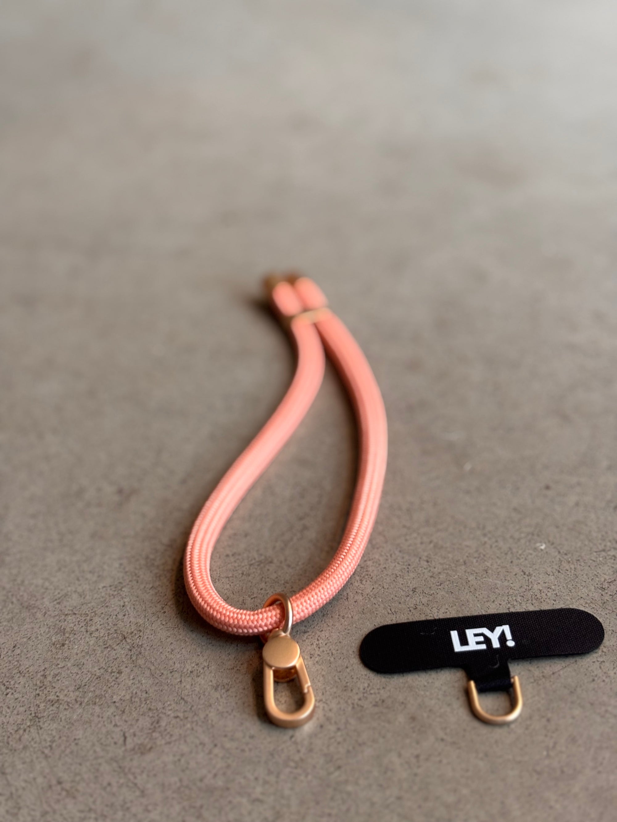 Ein pfirsichfarbener Schlüsselanhänger von LEY! mit goldener Schließe ruht auf einer grauen Oberfläche, neben einem schwarzen "LEY!"-Anhänger und einem kleinen goldenen Ring - perfekt, um Ihre Schlüssel oder Ihr Telefon stilvoll aufzubewahren.
