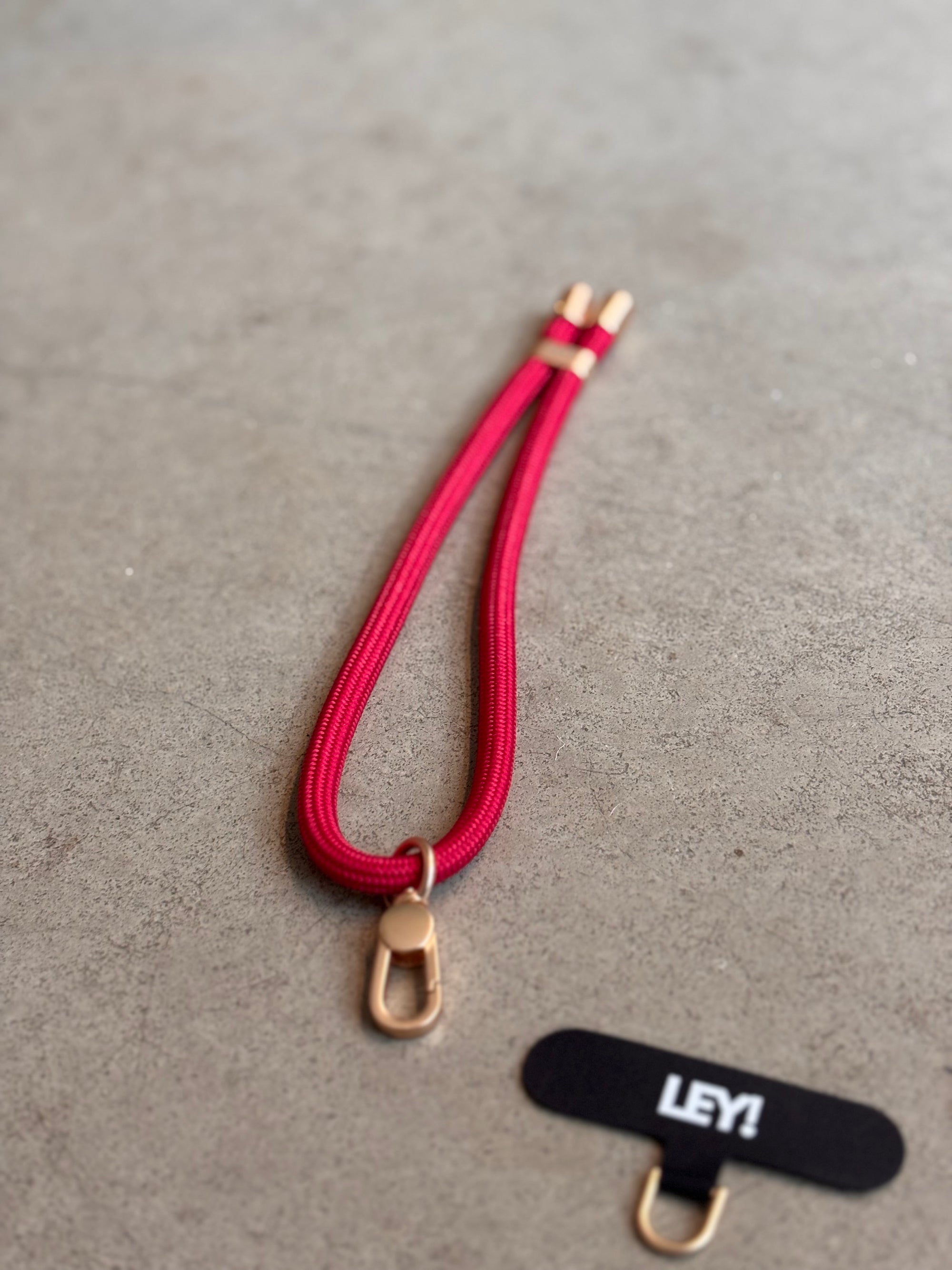 Ein roter Schlüsselanhänger von LEY! mit goldenen Clips liegt auf grauer Oberfläche, daneben ein kleines schwarzes Schild mit "LEY!" und goldenem Metallverschluss - ideal, um deinen Schlüssel immer griffbereit zu haben.