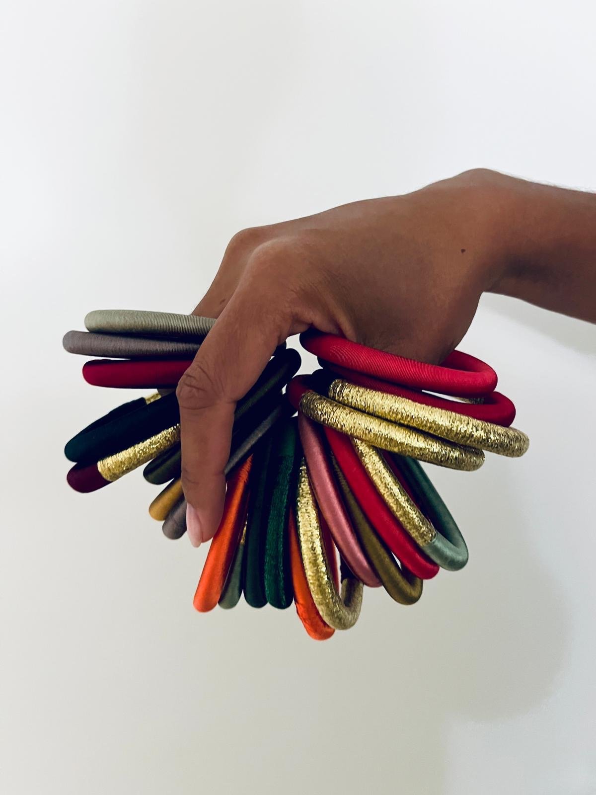 A hand hält mehrere bunte, ovale Armreife, darunter den handgefertigten "Armreif - mint" von LEY!, mit Garnwicklung im marokkanisch inspirierten Design, vor weißem Hintergrund.