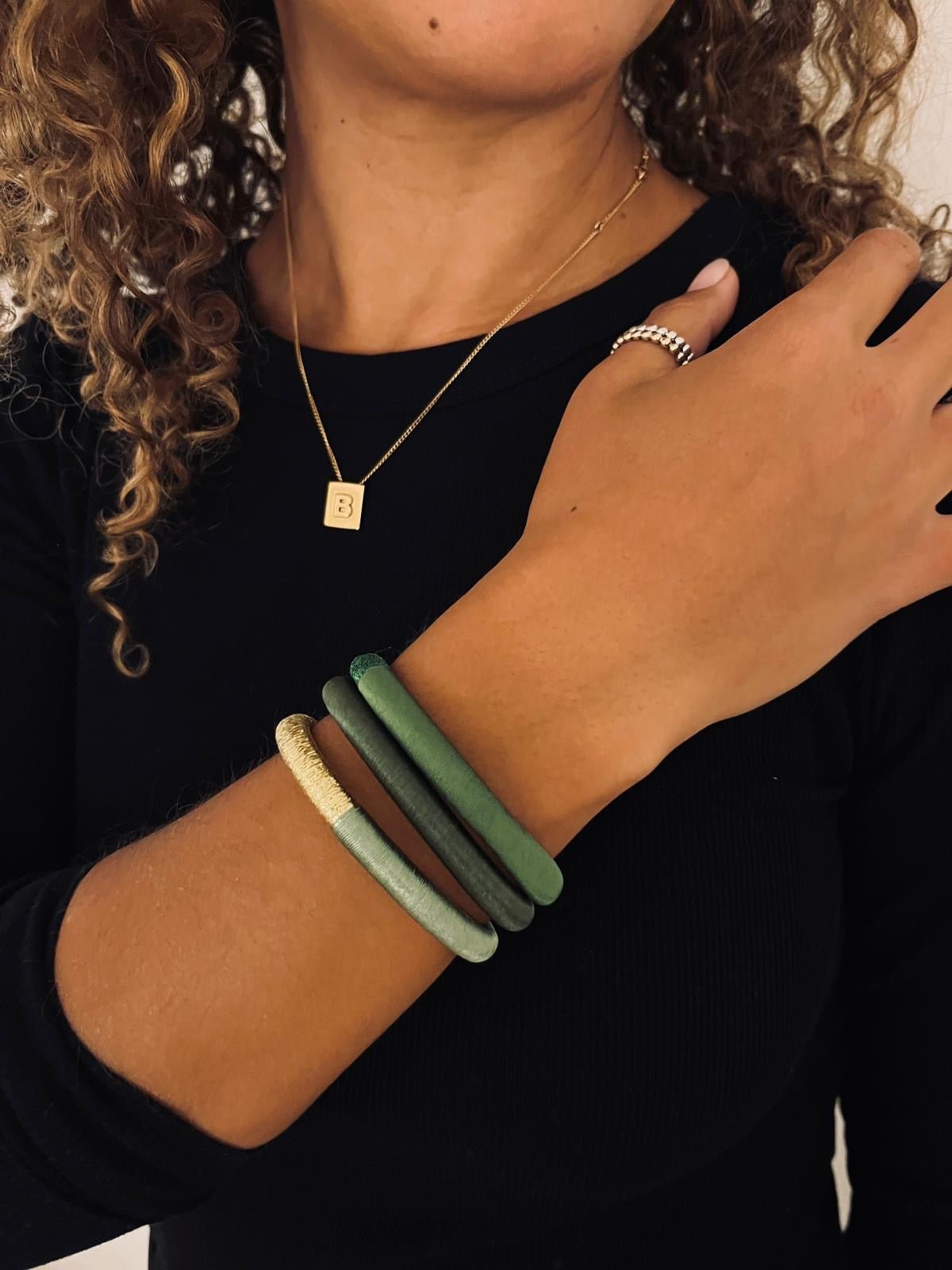 Eine Frau mit lockigem Haar trägt ein schwarzes Oberteil, eine goldene Halskette mit einem quadratischen "B"-Anhänger, einen Perlenring und drei klobige fliederfarbene LEY! Armreife am Handgelenk. Sie hält ihre Hand nahe der Schulter.