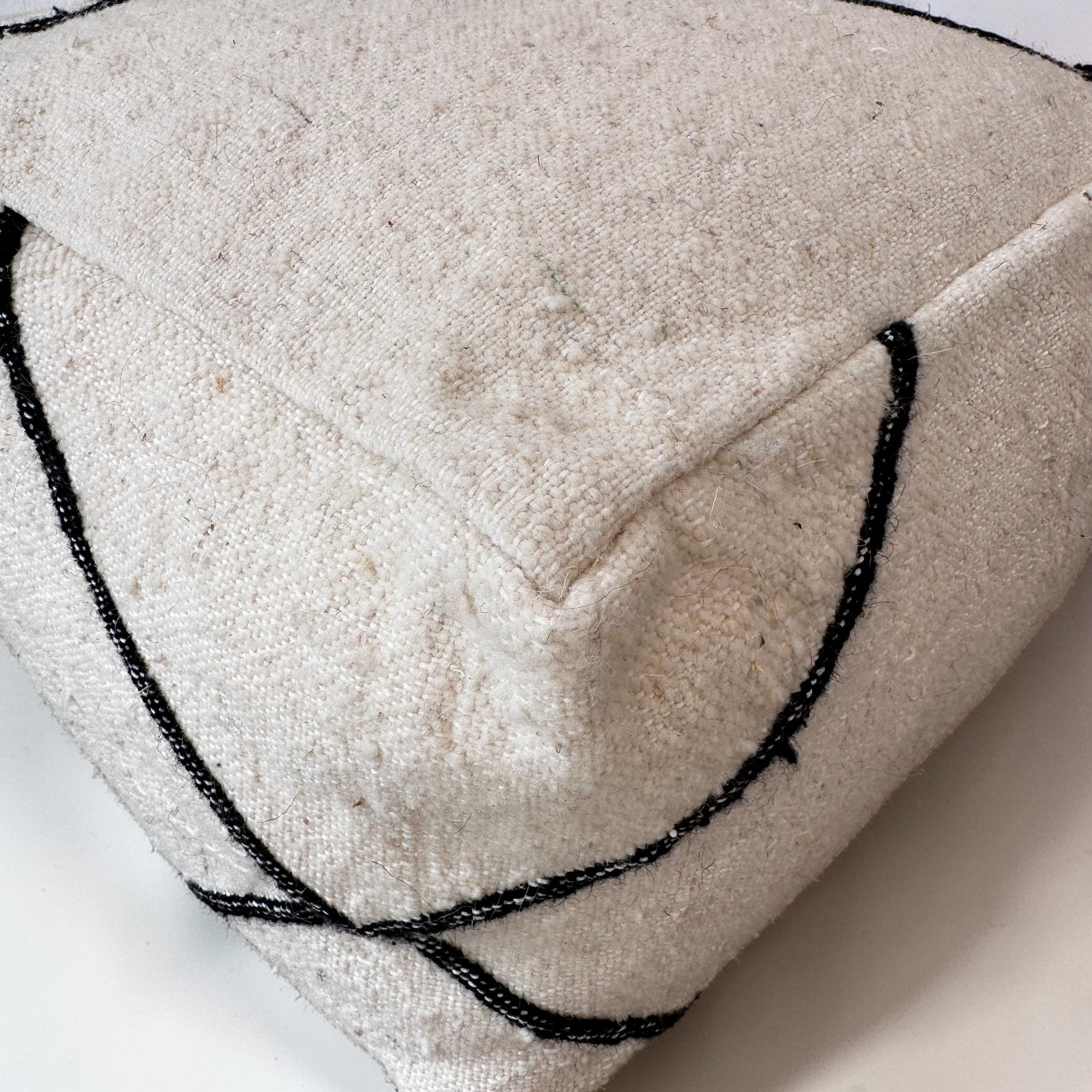 Nahaufnahme eines LEY! Kleines Bodenkissen - Meli Mini, handgefertigt aus off-white Schurwolle mit schwarzen gestickten Linien im minimalistischen abstrakten Muster. Ecke und Nähte sind sichtbar.