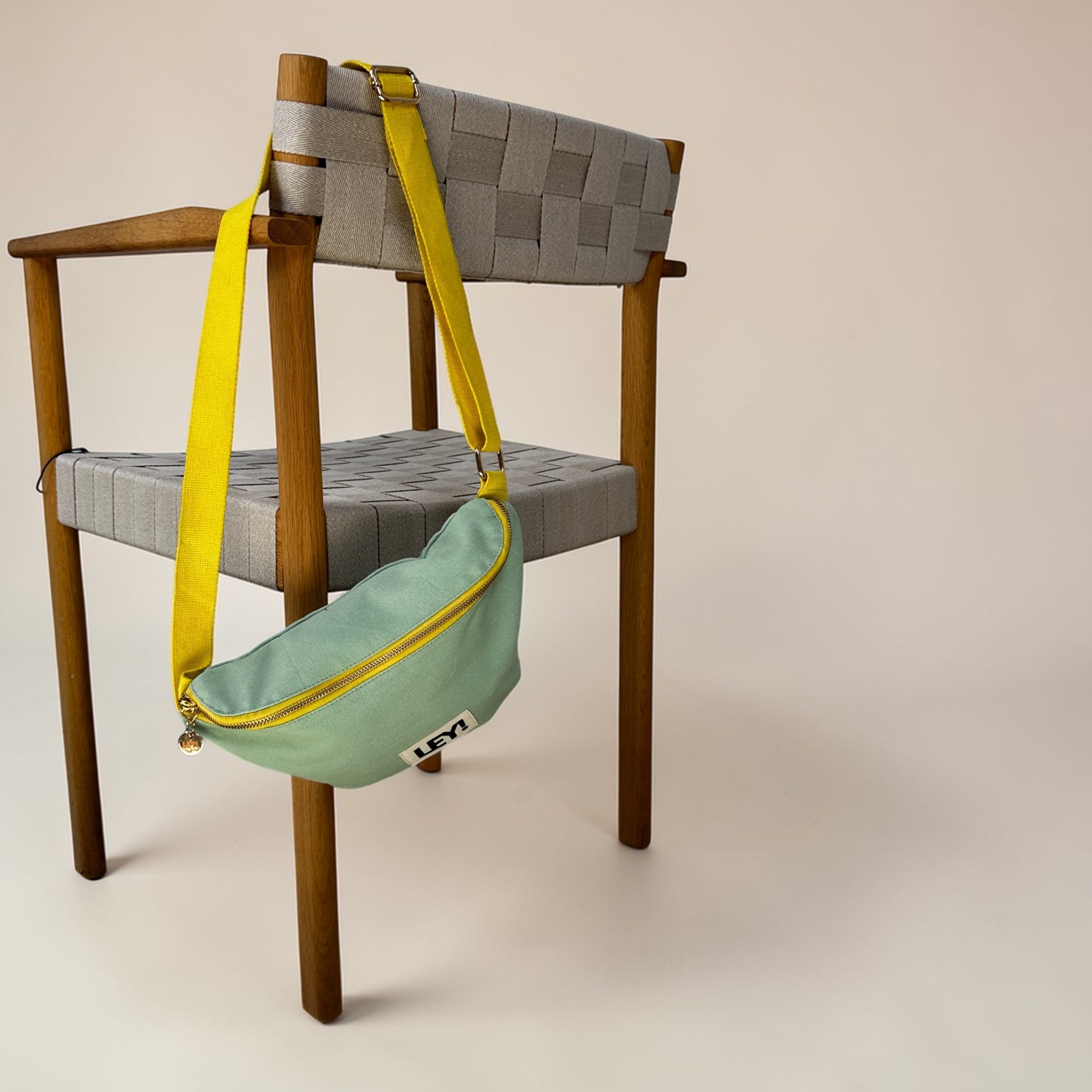 Die LEY! Bauchtasche - MINT-GELB von LEY! besteht aus einem hellgrünen Korpus mit gelbem Gurt, der über einen modernen Holzstuhl mit grauem Sitz und Rückenlehne vor beigem Hintergrund drapiert ist - ideal für den Festival-Stil.