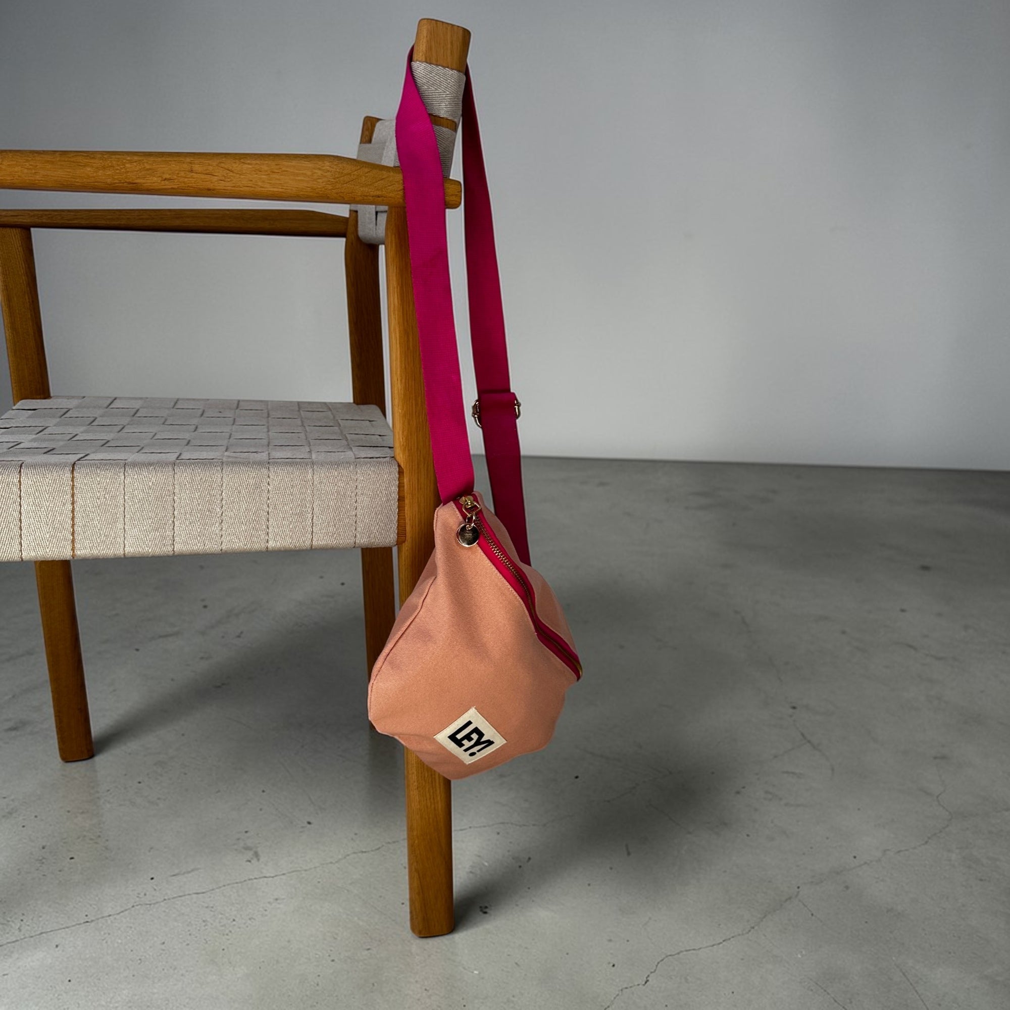 Die LEY! Bauchtasche - NUDE-PINK von LEY!, eine kleine pfirsichfarbene Tasche mit einem leuchtend pinken Riemen, hängt an der hölzernen Armlehne eines geflochtenen beigen Stuhls vor einem grauen Boden und hellgrauen Hintergrund - ideal für Ihr nächstes Festival.