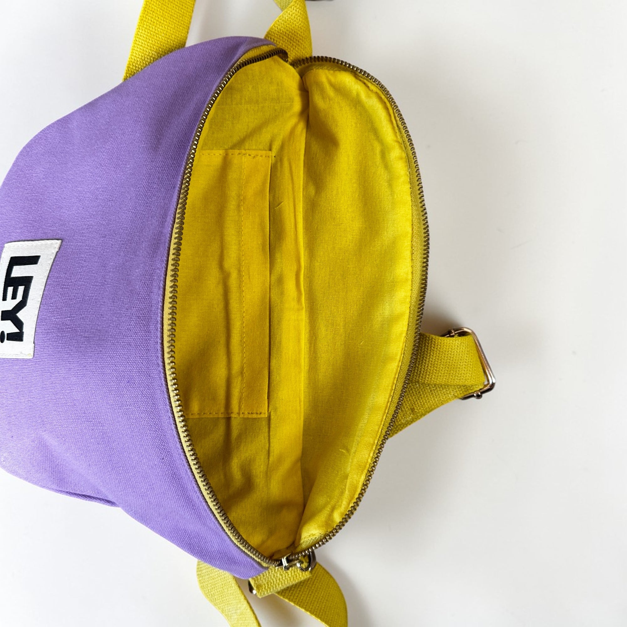Die LEY! Bauchtasche - FLIEDER-GELB ist eine kleine lilafarbene Tasche mit gelber Innenausstattung und Trageriemen, die durch Öffnen des Reißverschlusses ihre Innentasche und ein weißes Etikett mit schwarzem Text offenbart - ideal für Festivals.