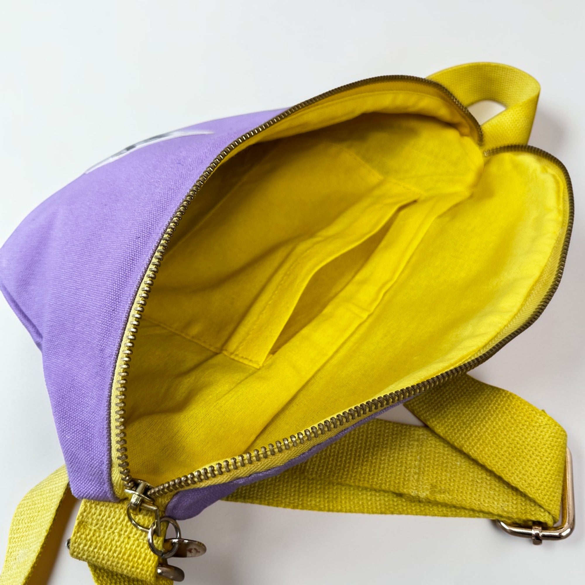 Die LEY! Bauchtasche - FLIEDER-GELB von LEY! zeigt sich in leuchtendem Lila mit gelbem Riemen. Nach dem Öffnen des Reißverschlusses kommen das leuchtend gelbe Innere und die Innentasche zum Vorschein - ein auffälliges Festival-Essential auf weißem Untergrund.