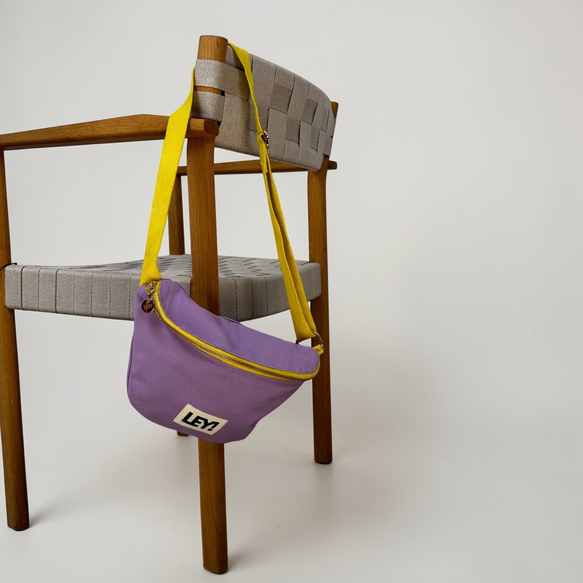 Ein Holzstuhl mit einer geflochtenen grauen Sitzfläche hat die LEY! Bauchtasche - FLIEDER-GELB über die Rückenlehne drapiert. Die lila-gelbe Tasche von LEY! hat einen weißen Aufnäher mit "LEY1" in schwarzer Schrift. Der Hintergrund ist einfarbig weiß.
