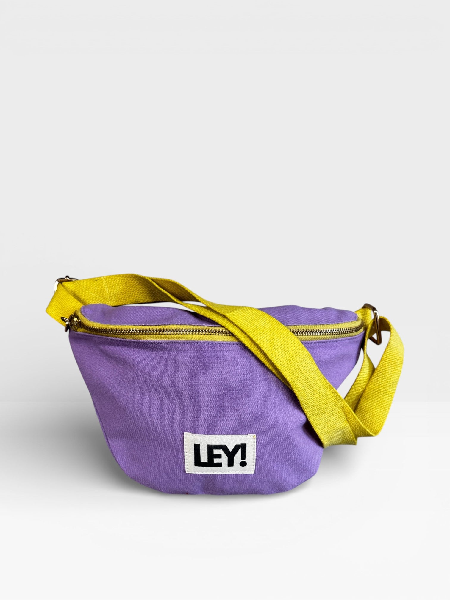 Die LEY! Bauchtasche - FLIEDER-GELB ist eine lilafarbene Stofftasche mit gelbem Riemen und Reißverschluss sowie einem weißen "LEY!"-Aufnäher auf der Vorderseite - ideal für deinen nächsten Festival-Look.