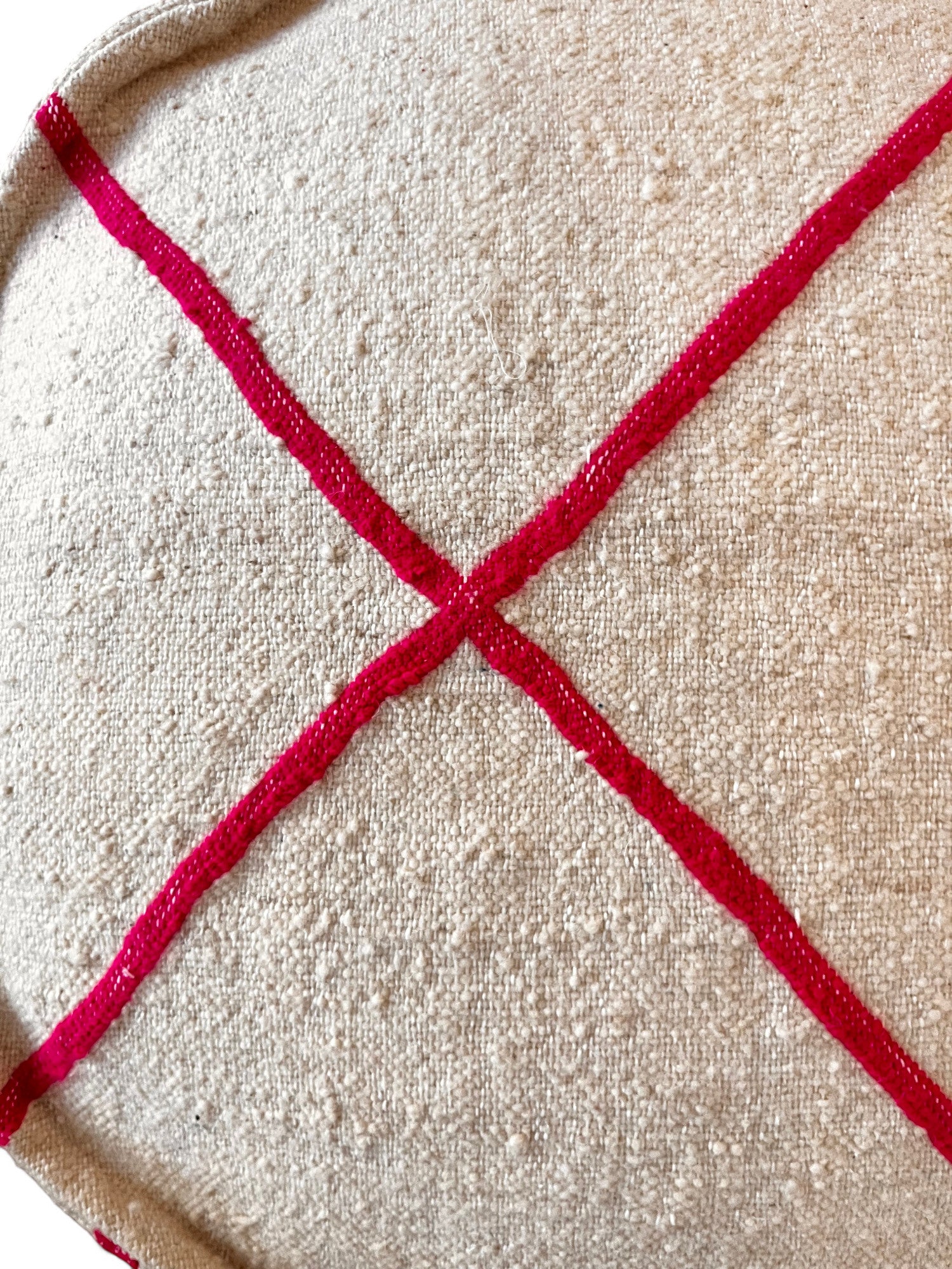 Eine Nahaufnahme des strukturierten beigen Stoffes des Bodenkissens Rund - Mina von LEY!, die zwei sich kreuzende rote Linien in Form eines "X" zeigt. Die genoppte Oberfläche suggeriert handwerkliche Qualität und macht dieses runde Kissen zu einem echten Unikat.