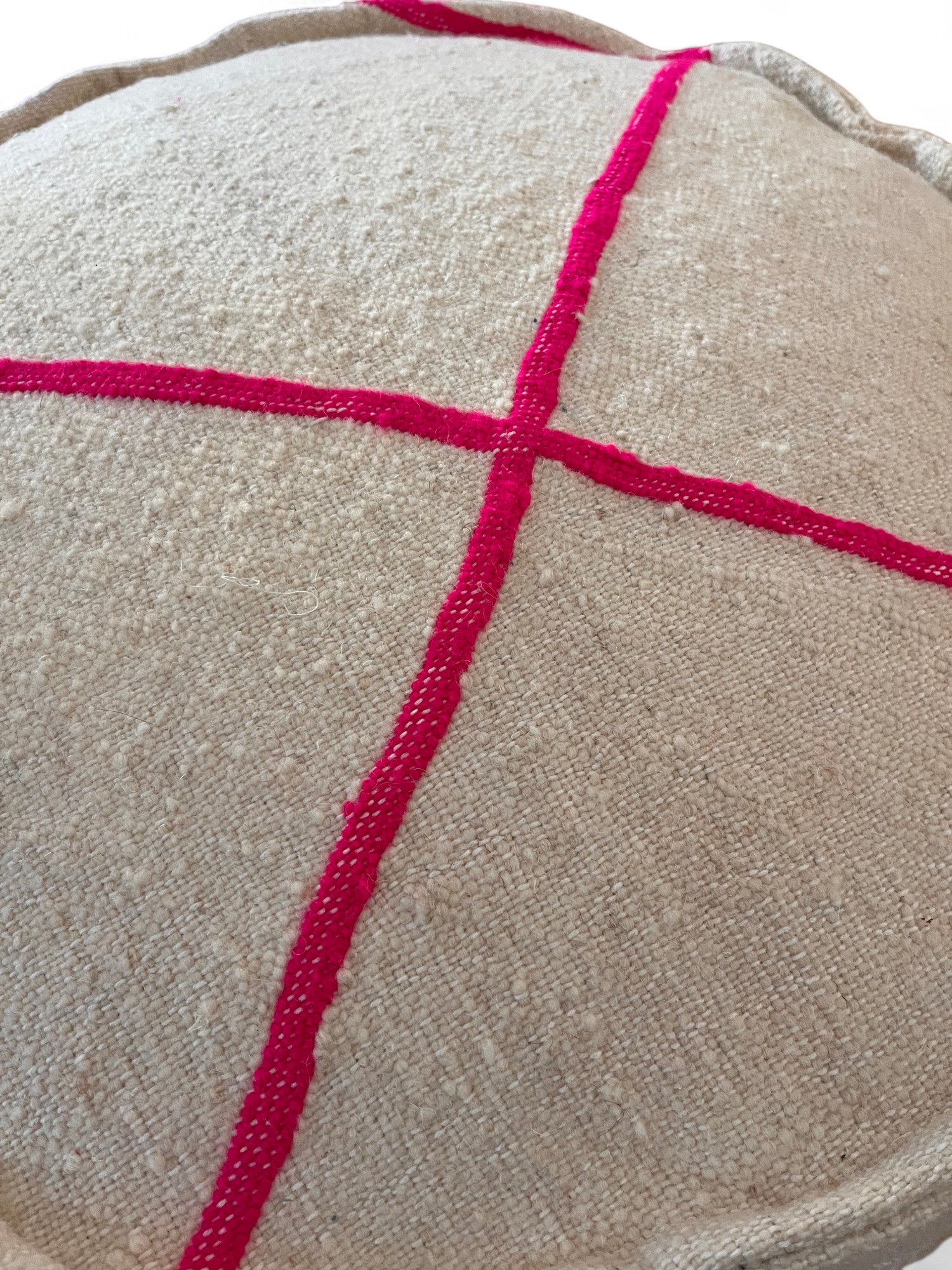Eine Nahaufnahme des LEY! Bodenkissen Rund - Mina zeigt seine beigefarbene Schurwolle-Oberfläche mit zwei leuchtend rosa gestickten Linien, die ein Kreuz bilden und detaillierte Handarbeit darstellen.