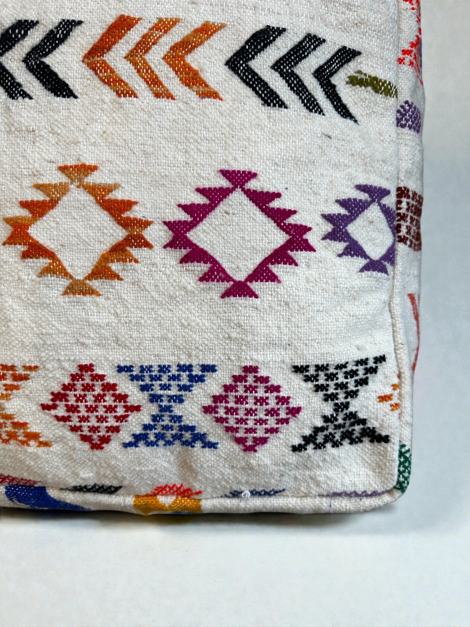 Eine Nahaufnahme des LEY! LEY Bodenkissen - Colorful Line - Simo, ein weißes handgefertigtes Bodenkissen mit lebhaften geometrischen Mustern - Diamanten und Chevrons - in orange, lila, schwarz, rot, blau und grüner Schurwolle.