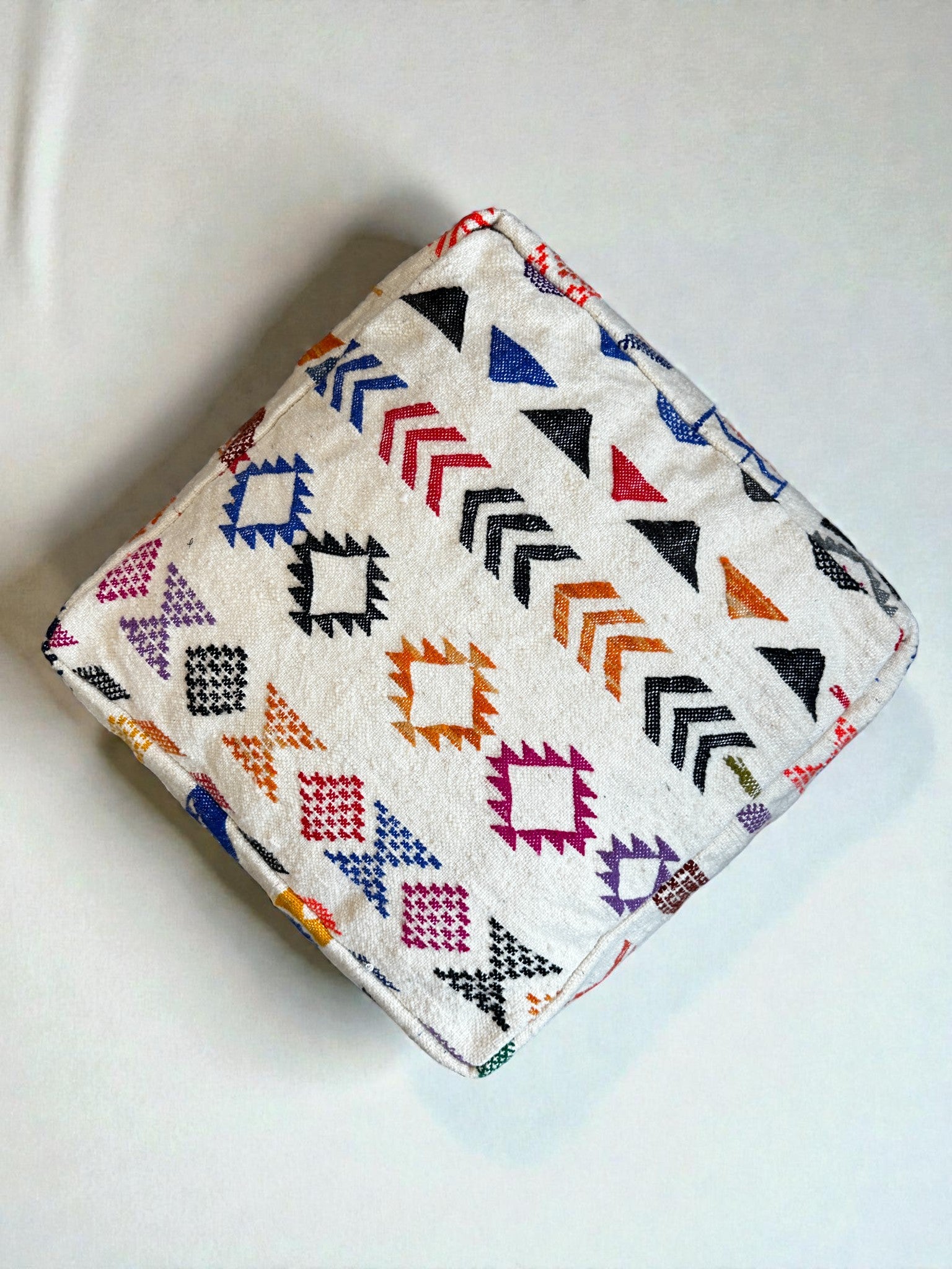Das LEY! LEY Bodenkissen - Colorful Line - Simo zeichnet sich durch ein weißes, quadratisches Design mit handgefertigten geometrischen und Pfeilmustern in Schwarz, Orange, Pink, Blau, Lila, Grün und Rot auf weicher Schurwolle auf einer glatten, hellen Oberfläche aus.