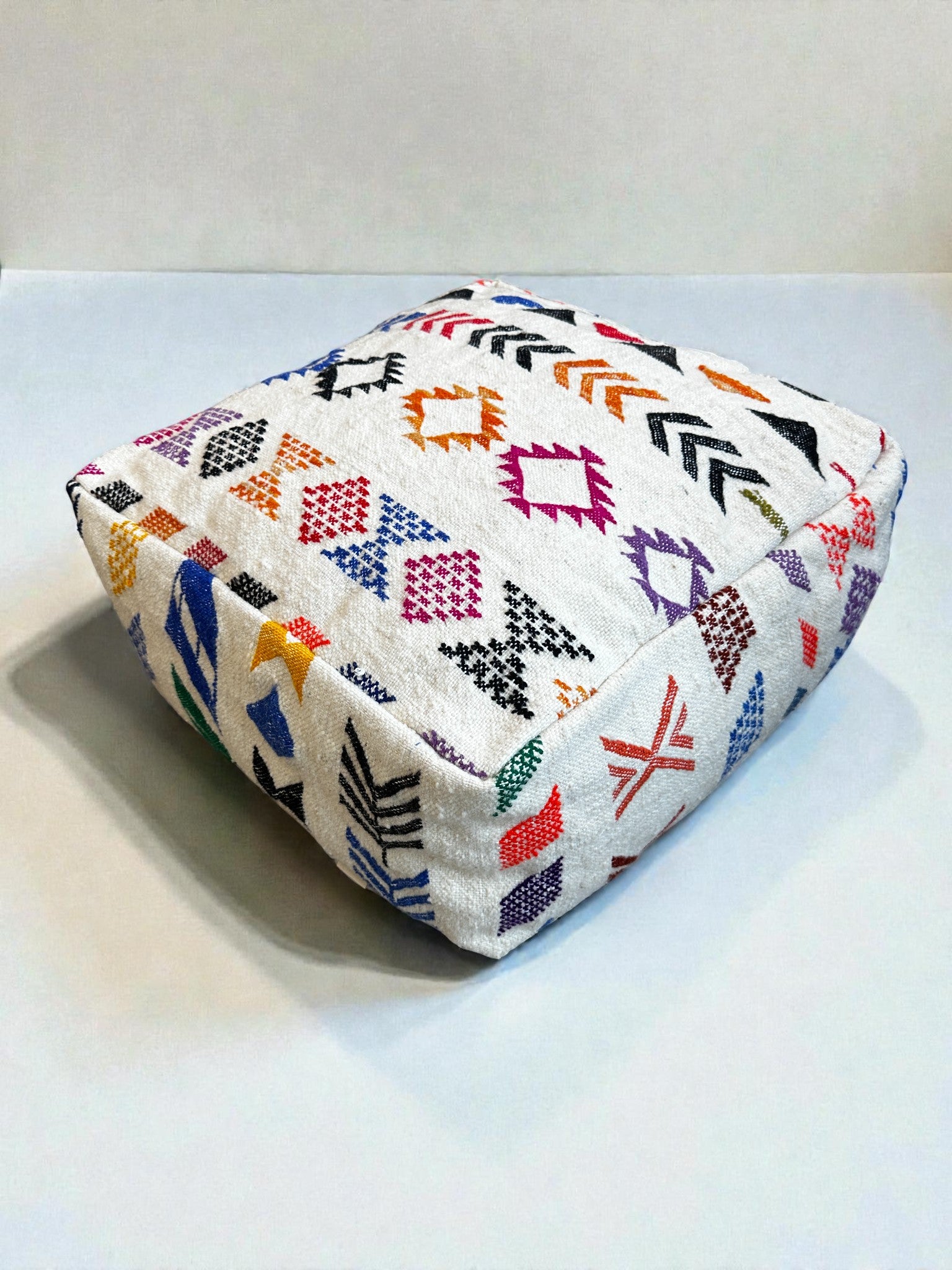 Das LEY! LEY Bodenkissen - Colorful Line - Simo ist ein quadratisches, weißes Bodenkissen aus Schurwolle, das in Handarbeit mit leuchtenden geometrischen Stickereien in Rot, Orange, Gelb, Lila, Grün und Schwarz auf einem unifarbenen, hellen Hintergrund hergestellt wird.