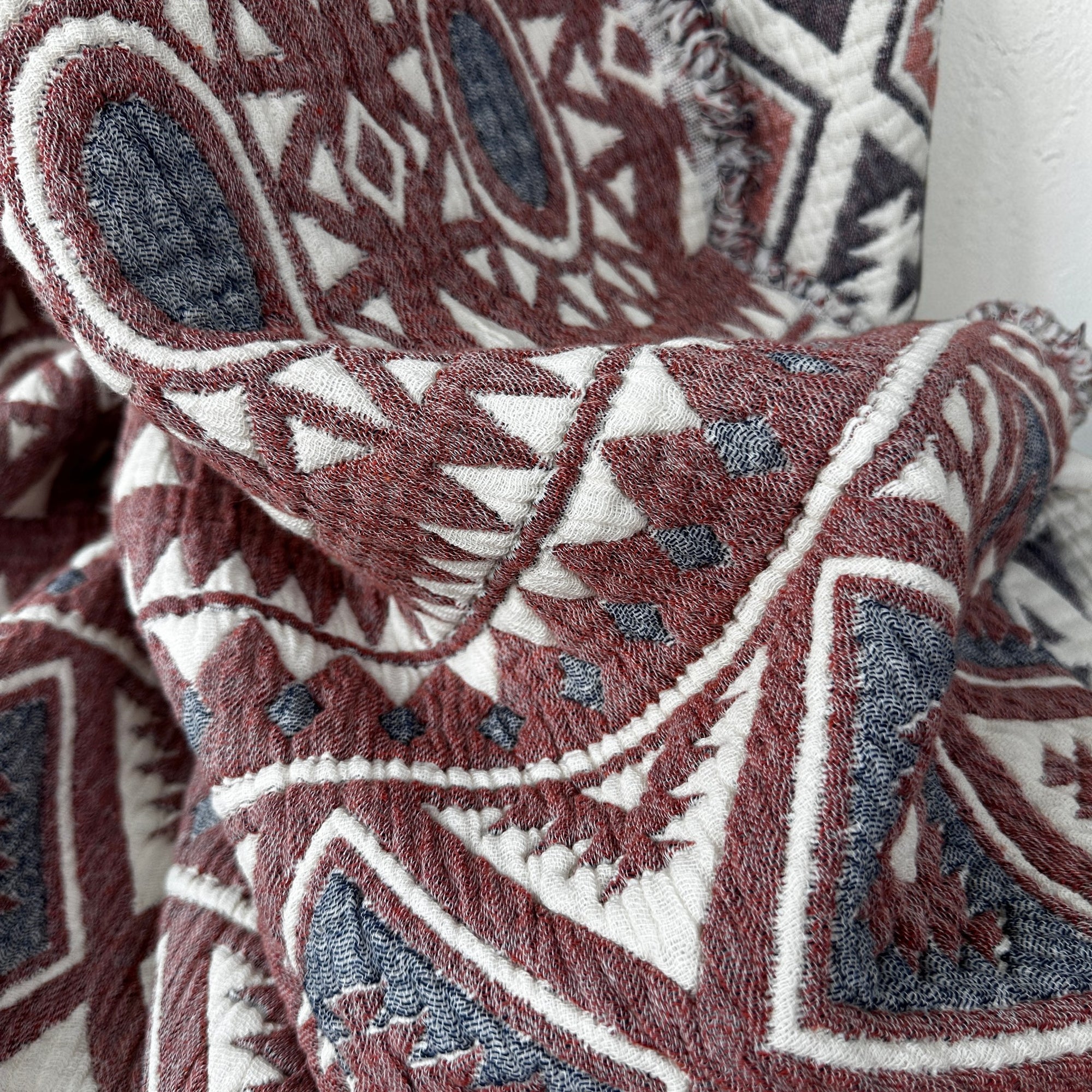 Nahaufnahme der CALMA Musselin Decke von LEY! (2,30 x 2,60 m) in Rot, Grau und Weiß. Das geometrische Boho-Design zeigt Rauten, Dreiecke und Ovale mit gestickten Akzenten und weichen, ausgefransten Kanten - perfekt für gemütlichen Komfort.