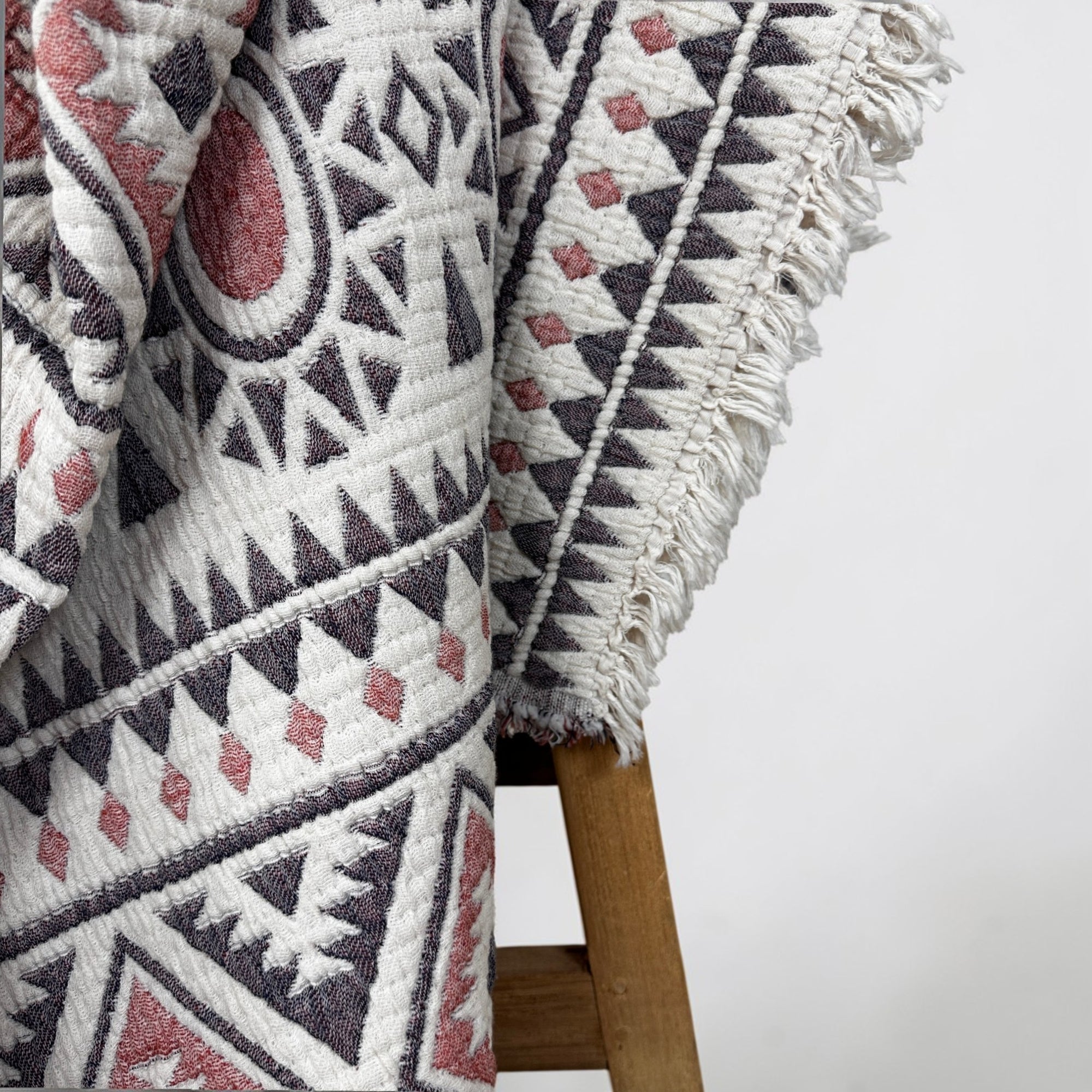 Eine Nahaufnahme der LEY! CALMA Musselin Decke in Rot-Grau-Weiß (2,30 x 2,60 m) zeigt ihr geometrisches Boho-Design und weiße Fransen, die über einen Holzstuhl drapiert sind, mit atmungsaktivem Stoff vor hellem Hintergrund.