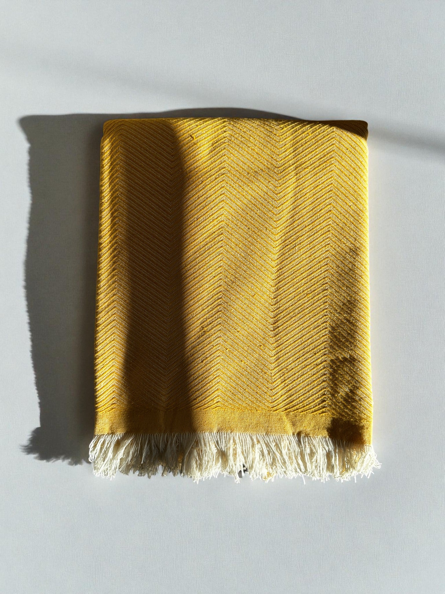Die ATLAS Decke - Leyla Line in Sonnengelb aus 100% Baumwolle mit dezentem Zickzack-Muster und weißen Fransen liegt sauber gefaltet auf einer hellen Fläche und wirft einen weichen Schatten nach links.