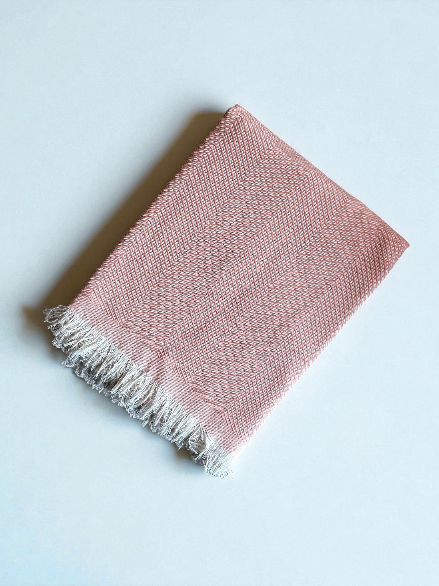 Die ATLAS Decke - Leyla Line - rosa ist eine gefaltete rosa Decke mit dezentem Fischgrätenmuster und weißen Fransen, ideal als Tagesdecke aus Baumwolle, auf einem schlichten hellen Hintergrund.