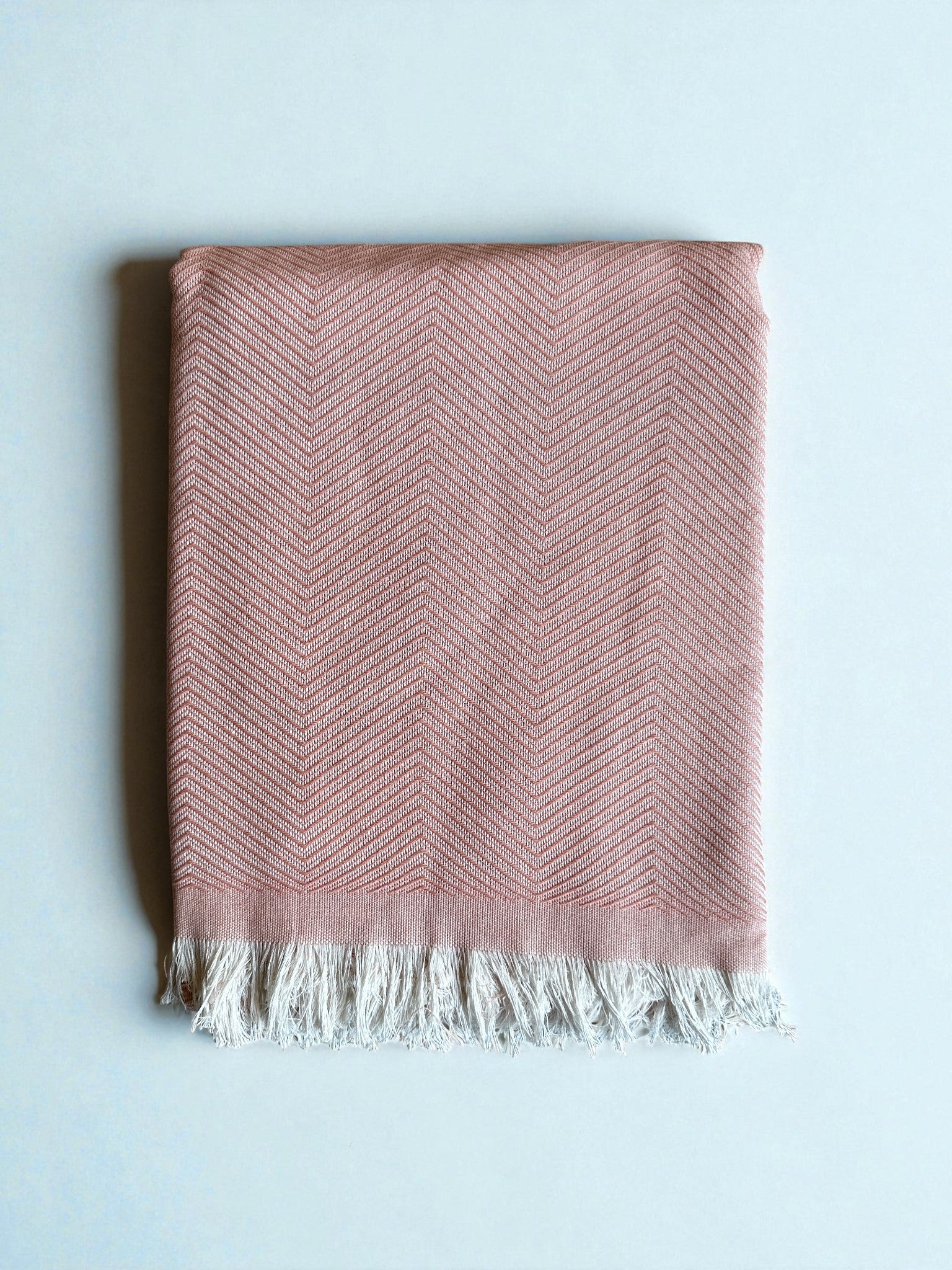 Die ATLAS Decke - Leyla Line - rosa ist eine rosafarbene Baumwolldecke mit Fischgrätmuster und weißen Fransen an einer Kante, die auf einer hellen Oberfläche gefaltet ist.