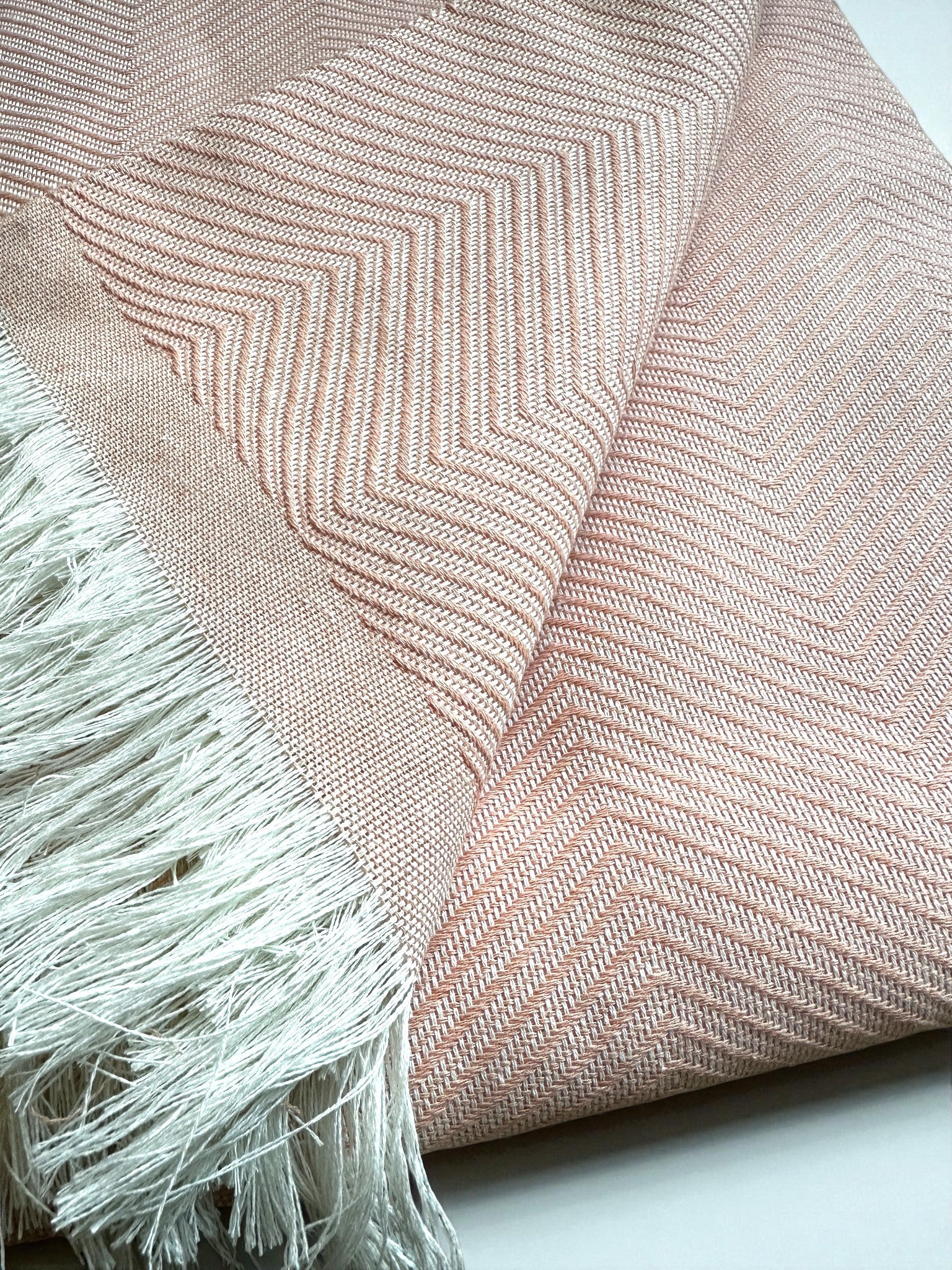 Die ATLAS Decke "Leyla Line" in rosa ist eine gefaltete, hellrosa gewebte Decke mit dezentem Fischgrätmuster und weißen Fransen an den Rändern, perfekt als Sofadecke oder Picknickdecke. Ausgestellt auf einer glatten Fläche.