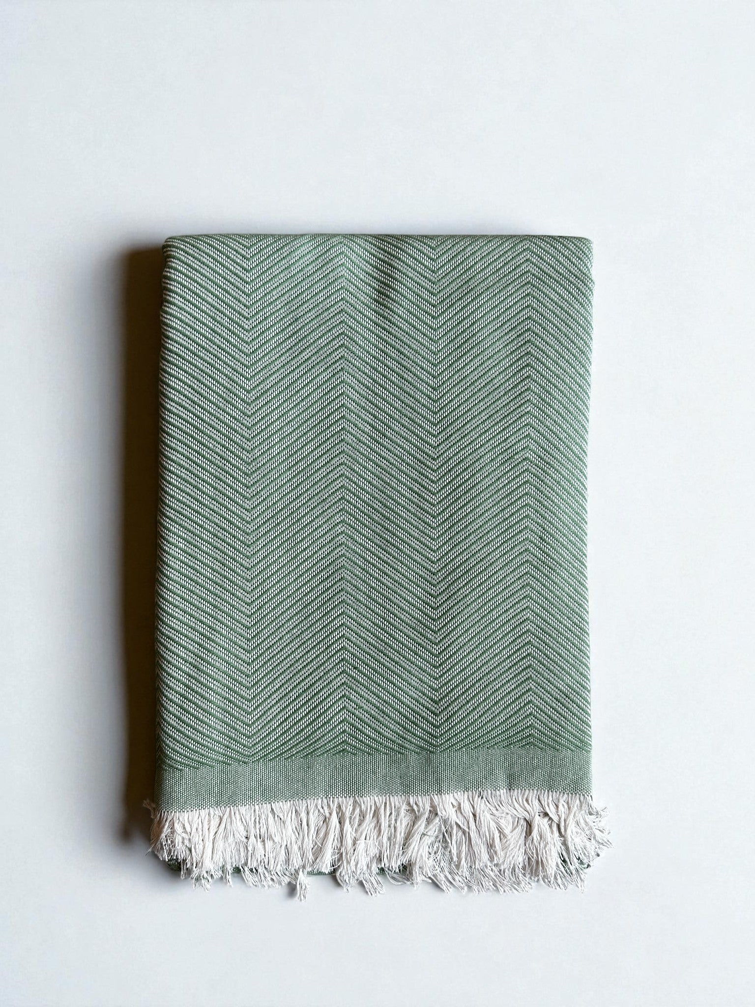 Die ATLAS Decke - Leyla Line - grün ist eine gefaltete, hellgrüne Baumwolldecke mit dezentem Chevron-Muster und weißen Fransen an einer Kante, die auf einem unifarbenen, hellen Hintergrund angebracht ist.