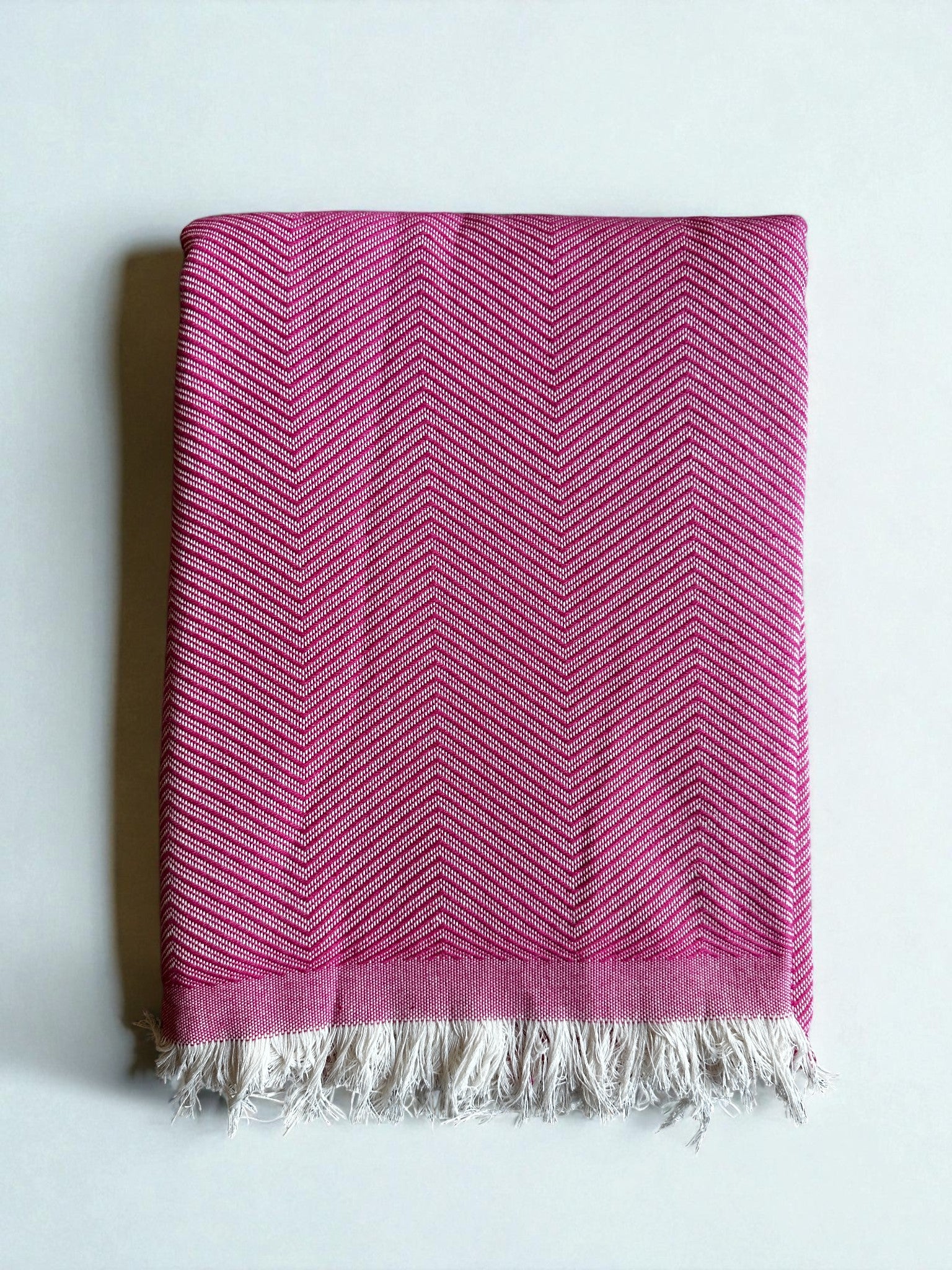 Eine fein säuberlich gefaltete ATLAS Decke - Leyla Line - rosa mit dezentem Fischgrätenmuster und weißen Fransen an den Rändern, dargestellt auf einem schlichten weißen Hintergrund.