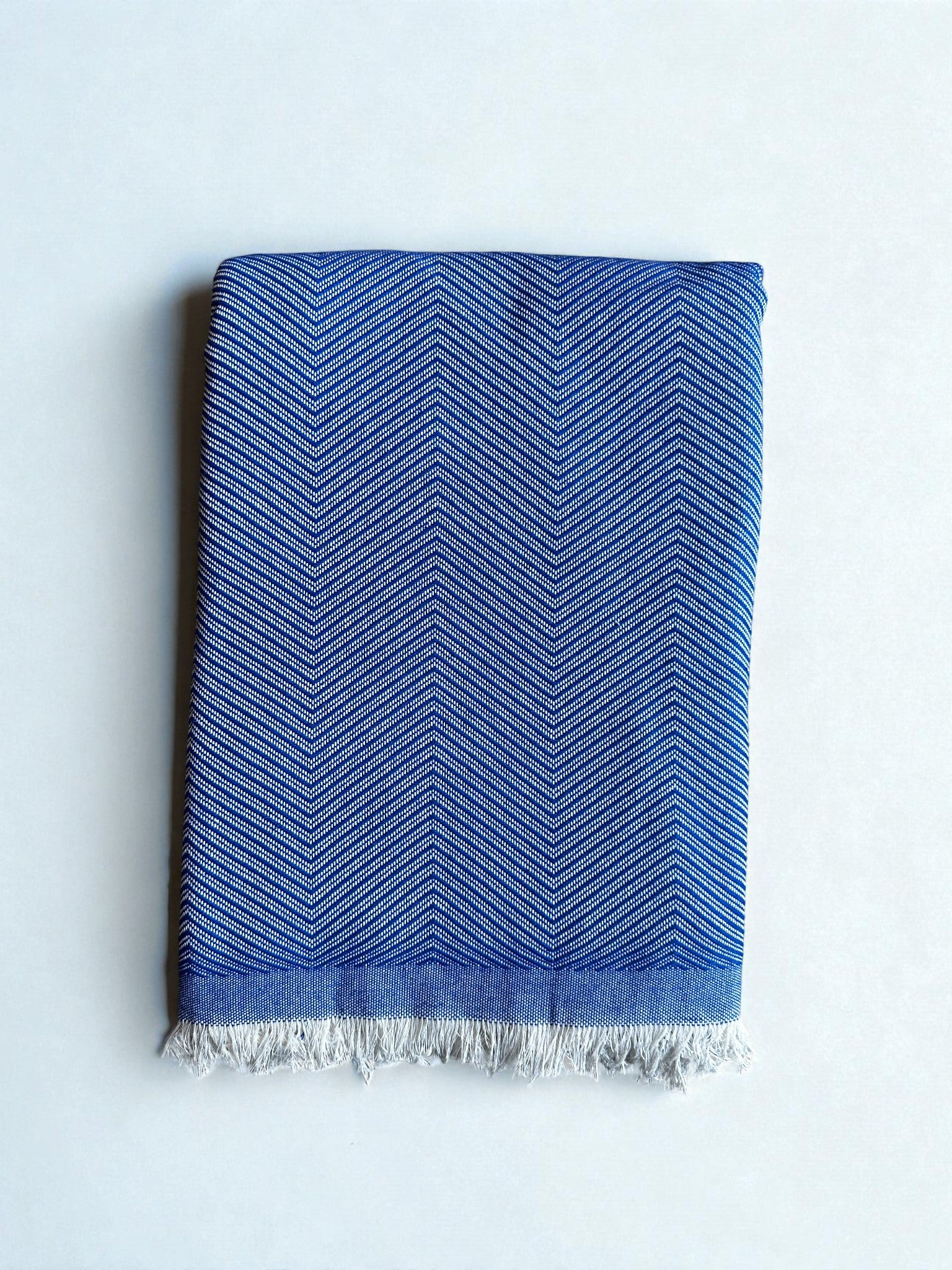 Die ATLAS Decke - Leyla Line - blau zeigt ein dezentes Chevron-Muster aus weicher Baumwolle, sauber gefaltet mit einem kurzen weißen Fransenrand an einer Kante und auf hellem Grund.