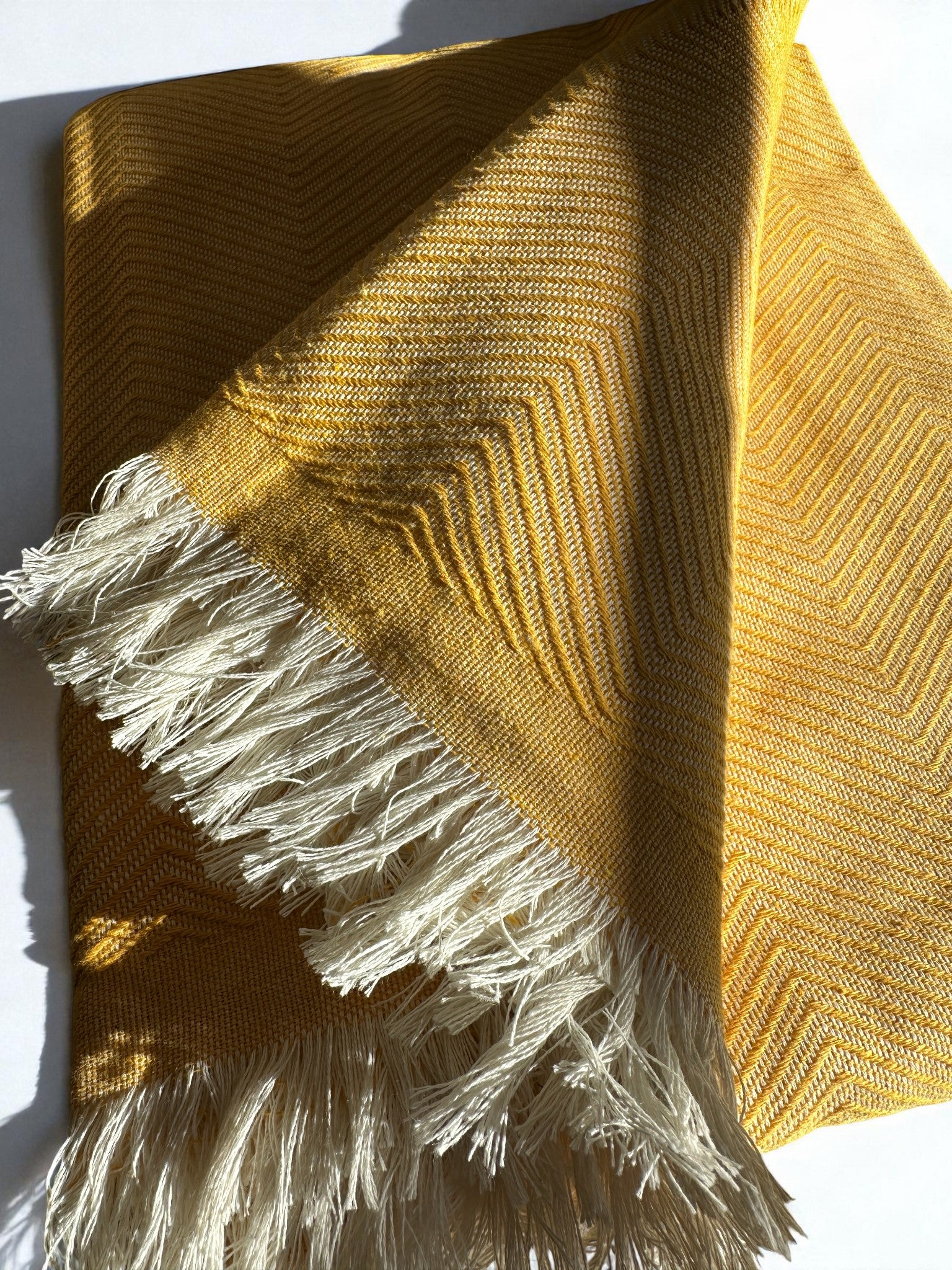 Die ATLAS Decke - Leyla Line in Sonnengelb zeichnet sich durch ein dezentes Chevron-Muster, weiße Fransen und strukturierte Baumwolle aus. Das Sonnenlicht hebt die minimalistische Webart und die Fransen dieser eleganten Decke hervor.