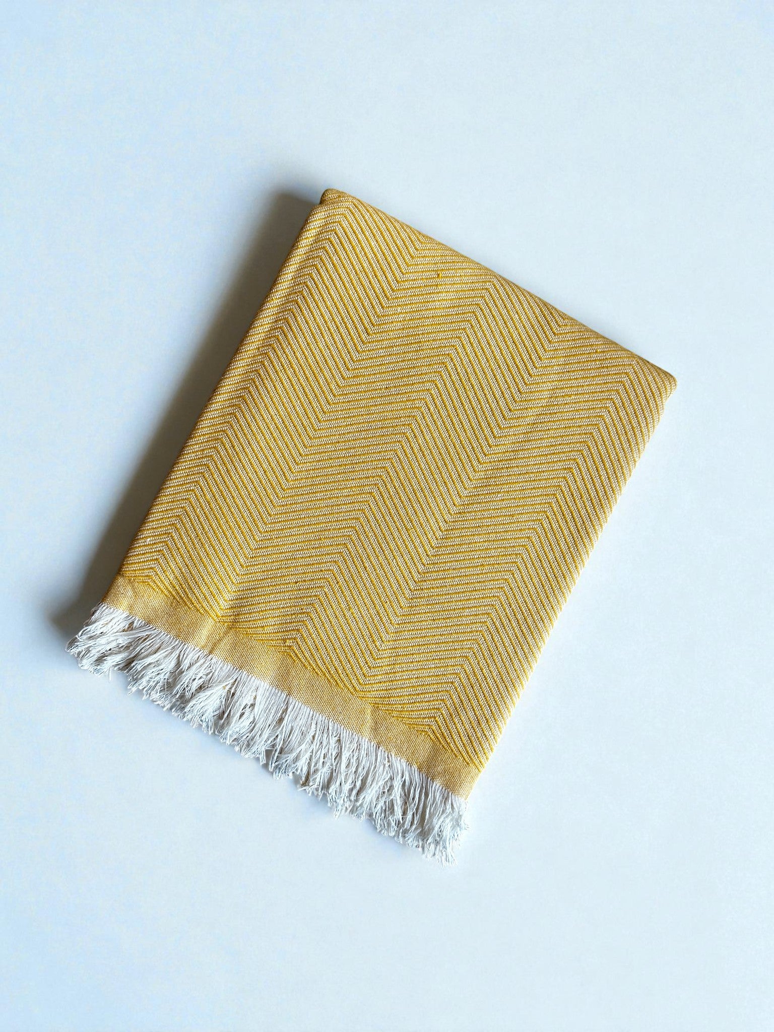 Die ATLAS Decke - Leyla Line - Sonnengelb, eine gelbe gewebte Decke mit Chevron-Muster und weißen Fransen, ruht auf einer hellen Oberfläche - ideal als minimalistische Tagesdecke Baumwolle für moderne Innenräume.