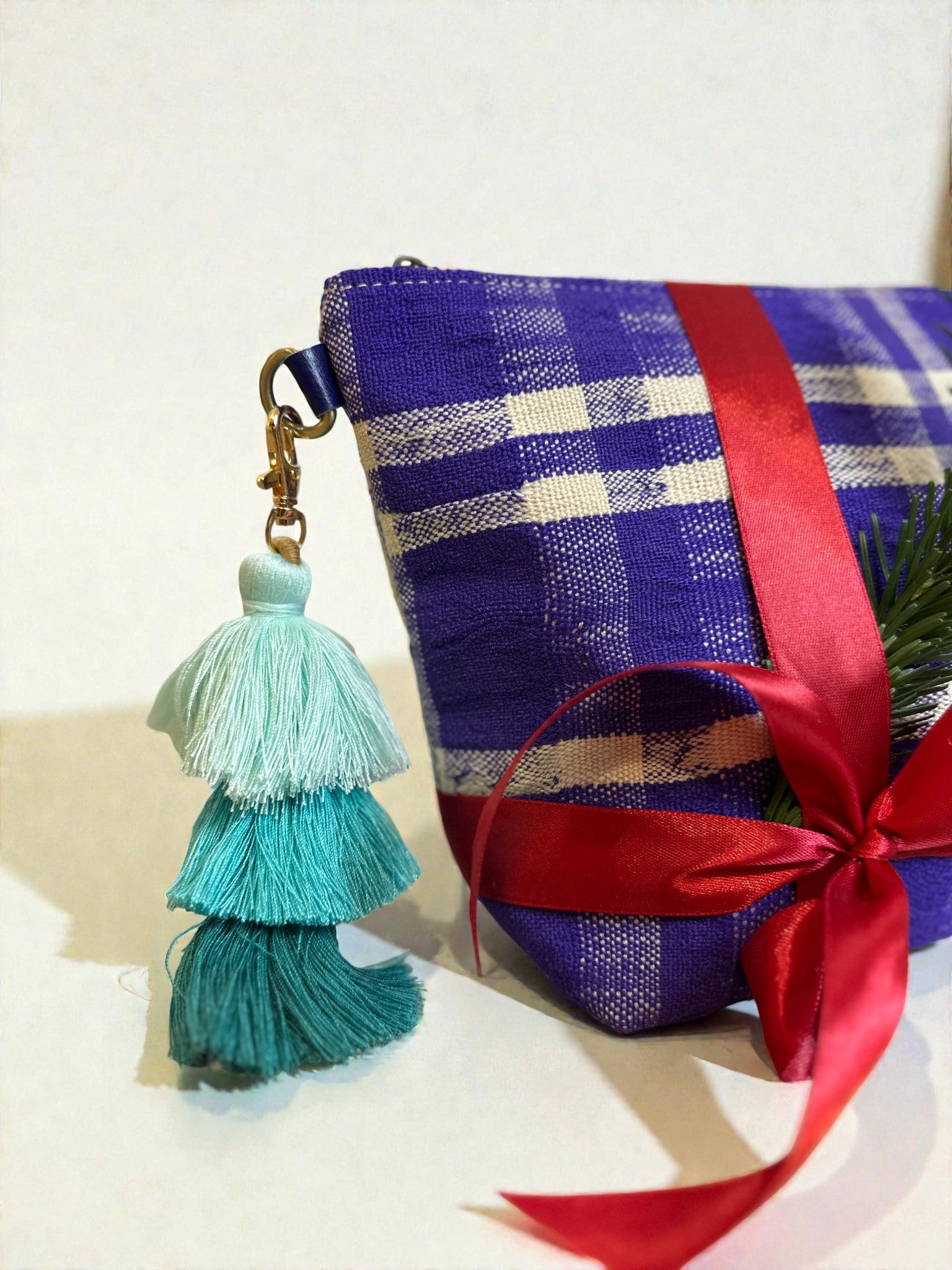 Abgebildet ist ein lila-weiß kariertes Täschchen mit roter Schleife und dem LEY! Schlüsselanhänger QUASTE in Blau- und Grüntönen sowie einem kleinen grünen Zweig - eine ideale Accessoire-Geschenkidee.