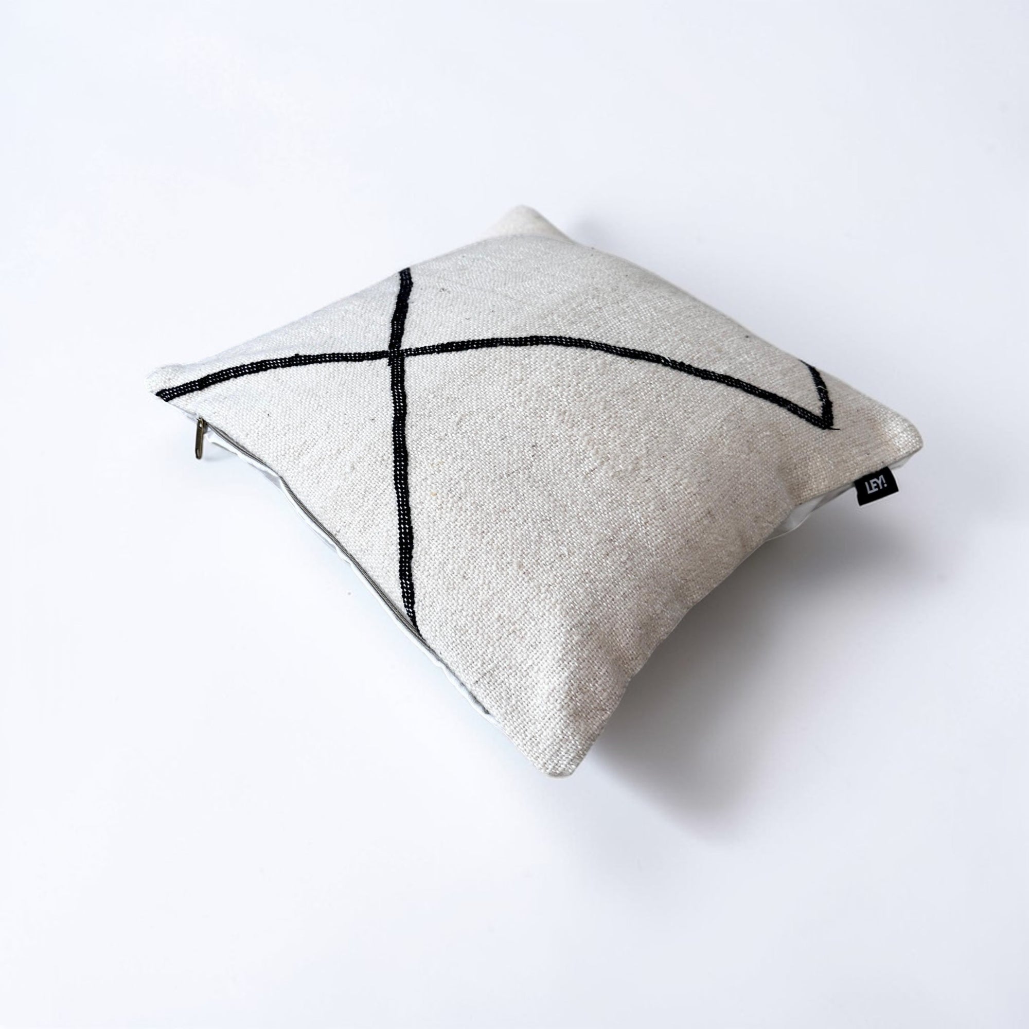 Das LEY! Kissen 50 x 50 - Meli ist ein quadratisches, beigefarbenes Schurwolle-Kissen mit einem einfachen, schwarzen, geometrischen Linienmuster und einem kleinen, schwarzen Anhänger an einem Rand, der auf einem schlichten, weißen Hintergrund angebracht ist.