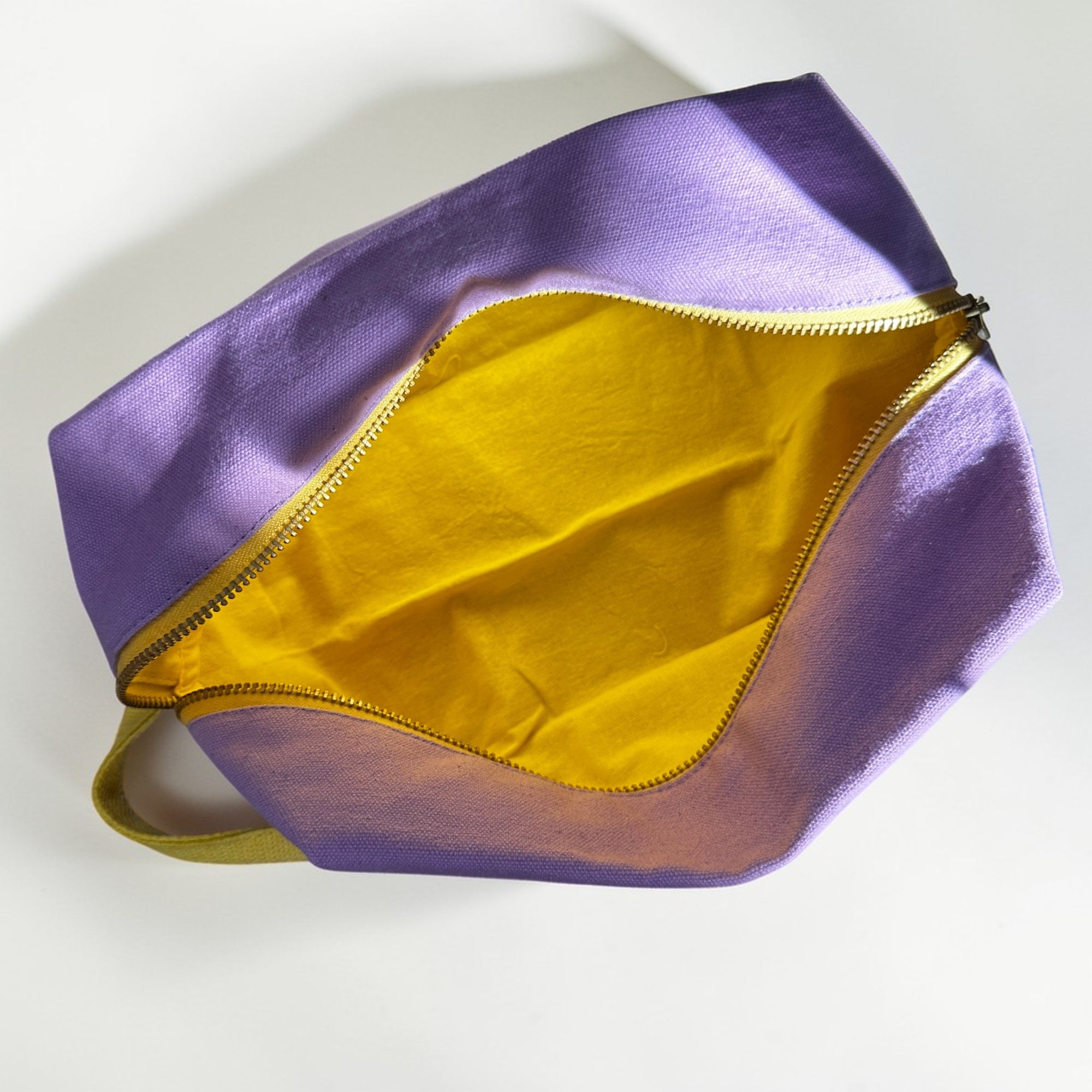 Der LEY! Pouch - FLIEDER-GELB von LEY! ist mit seinem lilafarbenen Äußeren, dem gelben Inneren und dem goldenen Reißverschluss - teilweise von oben geöffnet auf weißem Hintergrund - ein ideales Accessoire für Kulturbeutel Reisen.