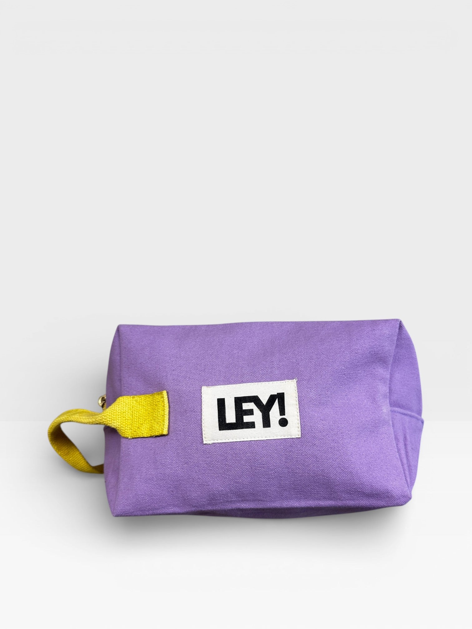 Der LEY! Pouch - FLIEDER-GELB von LEY! ist ein rechteckiger lavendelfarbener Beutel mit einem gelben Band und einem auffälligen "LEY!"-Aufnäher, ideal als Kulturbeutel Reisen oder Festival-Accessoire, abgebildet auf einem schlichten weißen Hintergrund.