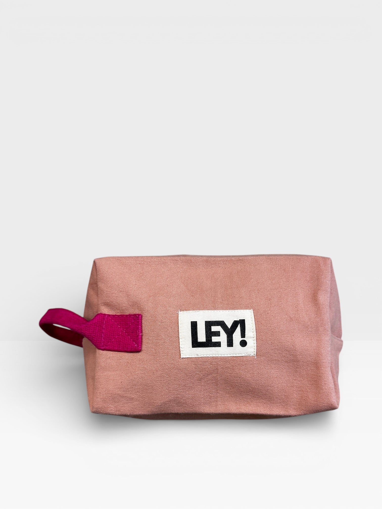 Die LEY! Pouch - NUDE-PINK von LEY! zeichnet sich durch einen rechteckigen, rosafarbenen Stoff, einen kastanienbraunen Henkel und einen weißen Aufnäher mit schwarzem "LEY!"-Schriftzug aus - ideal als stylisches Festival-Accessoire oder als Reisetasche, dargestellt auf einem schlichten weißen Hintergrund.