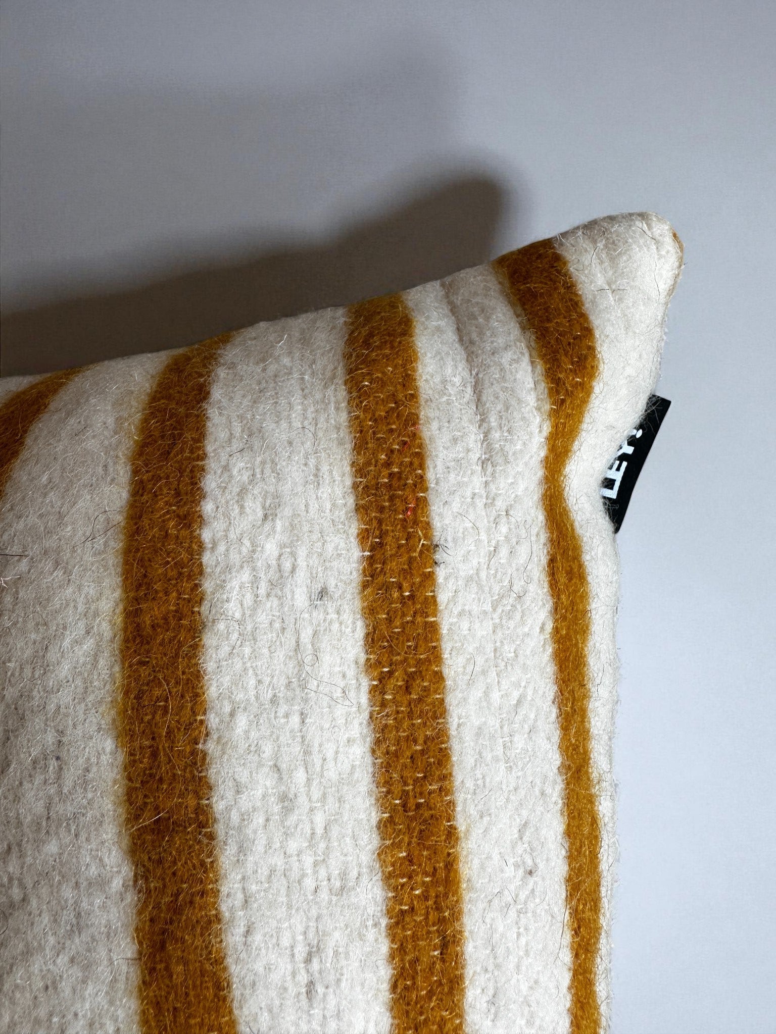 Eine Nahaufnahme des LEY! Kissen - WINTER EDITION - SONNE (30 x 50) zeigt die strukturierte Wolle und die kräftigen senfgelben und weißen Streifen, mit einem kleinen schwarzen Etikett am Saum. Der Hintergrund ist einfarbig und hell gefärbt.