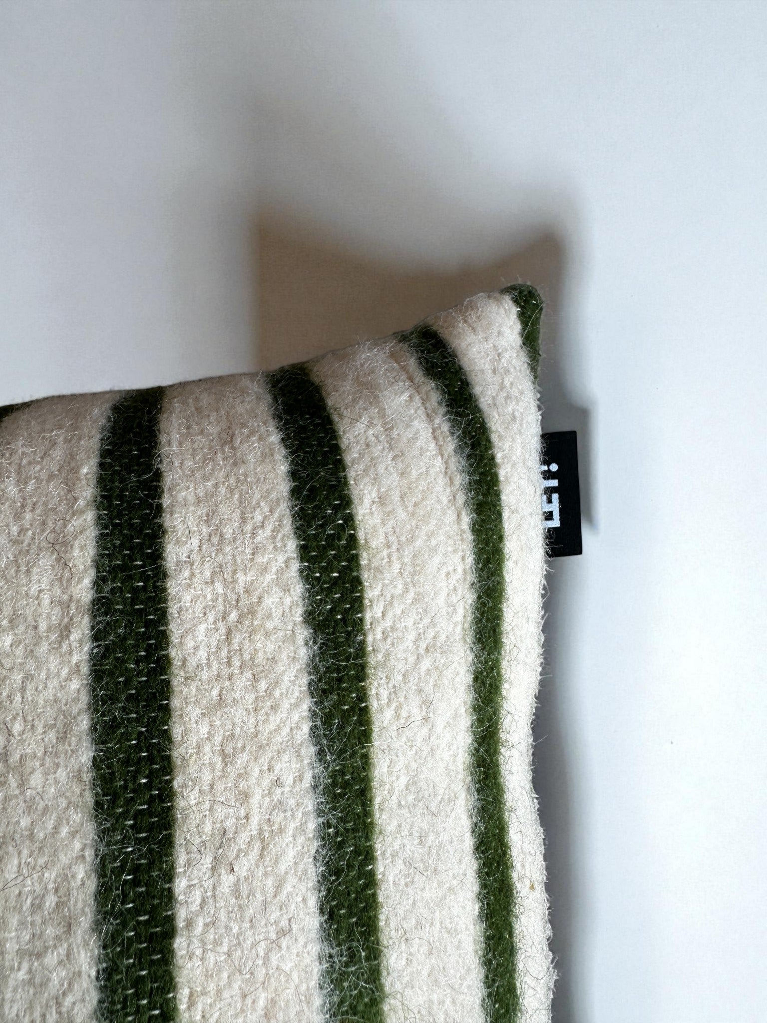 EIN LEY! Kissen - WINTER EDITION - TANNE (30 x 50), cremefarben mit dunkelgrünen vertikalen Streifen und einem schwarzen Stoffanhänger auf der rechten Seite, wird auf einer weißen Fläche ausgestellt.