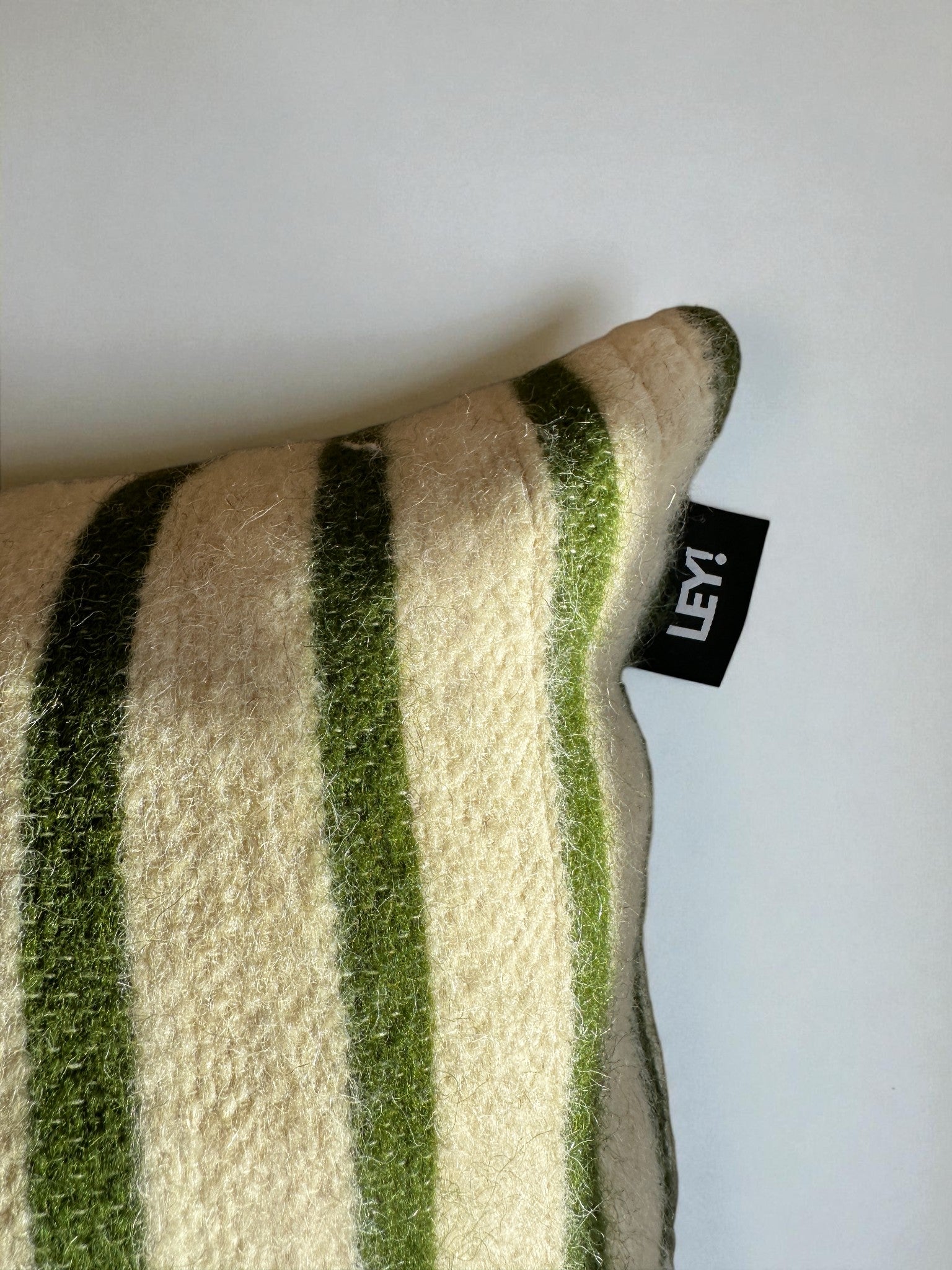 Eine Nahaufnahme des Kissens - WINTER EDITION - TANNE - 30 x 50 von LEY!, einem beigen Kissen mit grünen Streifen, zeigt an einer Ecke ein schwarzes LEY! Der Hintergrund ist eine glatte, helle Fläche.