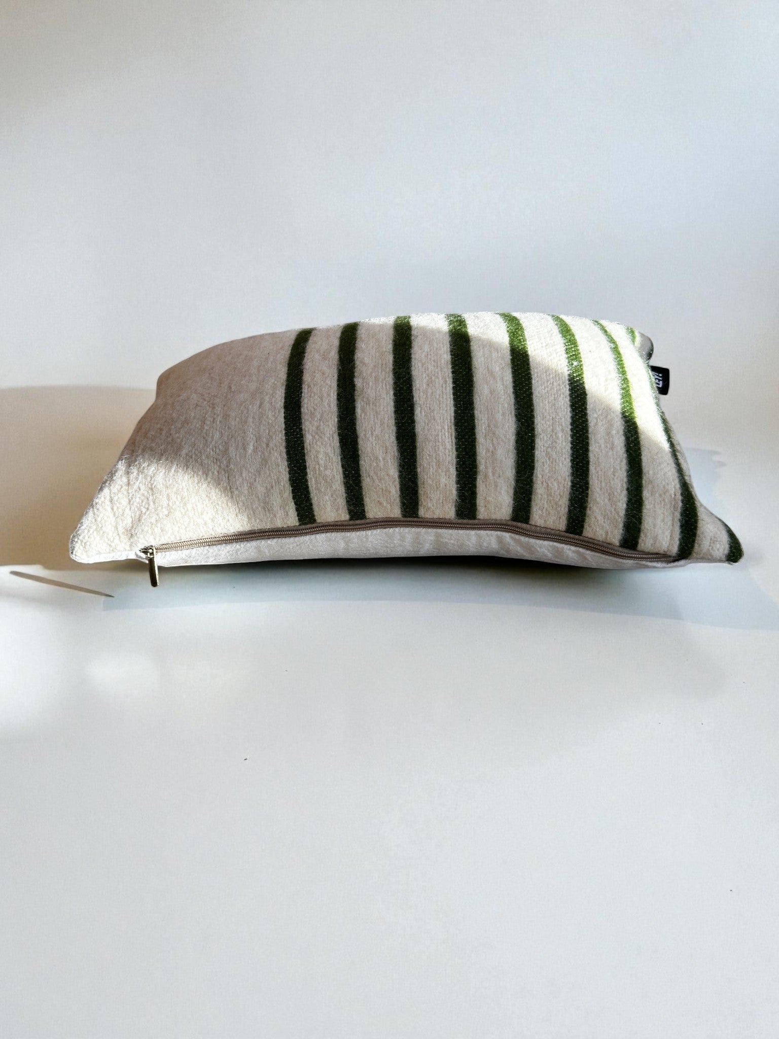 Das LEY! Kissen - WINTER EDITION - TANNE - 30 x 50 hat eine cremefarbene Basis mit dunkelgrünen Streifen, handgefertigt in Marrakesch, mit einem sichtbaren Reißverschluss an einer Kante und ruht auf einer weißen Oberfläche.