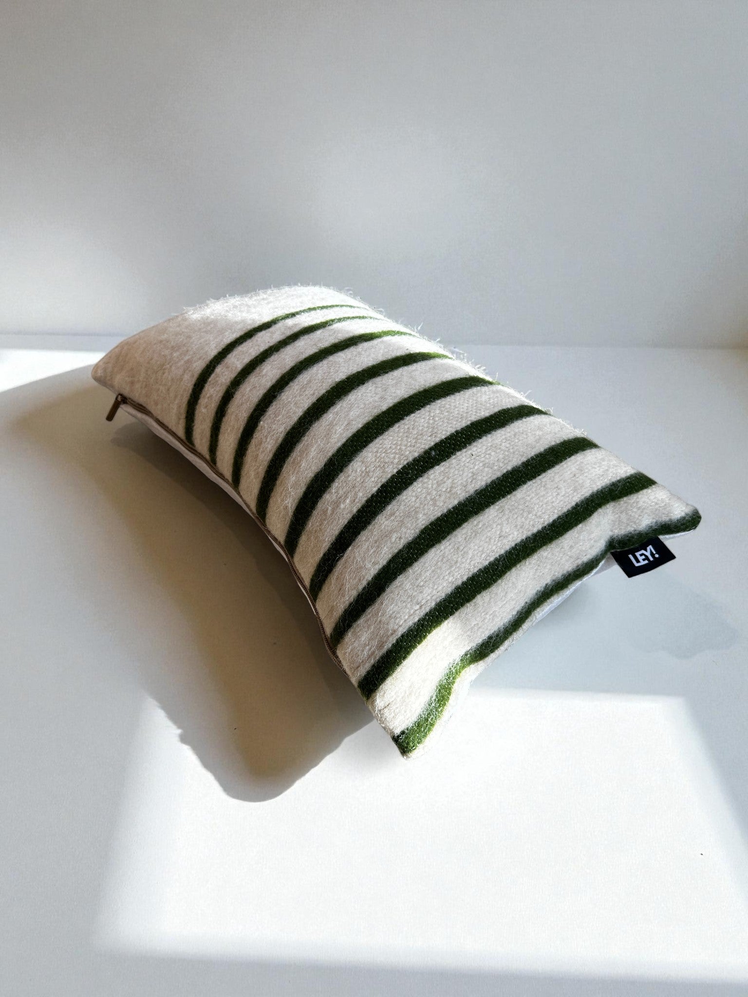 Ein rechteckiges LEY! Kissen - WINTER EDITION - TANNE - 30 x 50 mit grünen und weißen Streifen ruht auf einer weißen Fläche und hat ein schwarzes Seitenschild. Das Sonnenlicht wirft einen sanften Schatten und unterstreicht die gemütliche Winteratmosphäre.