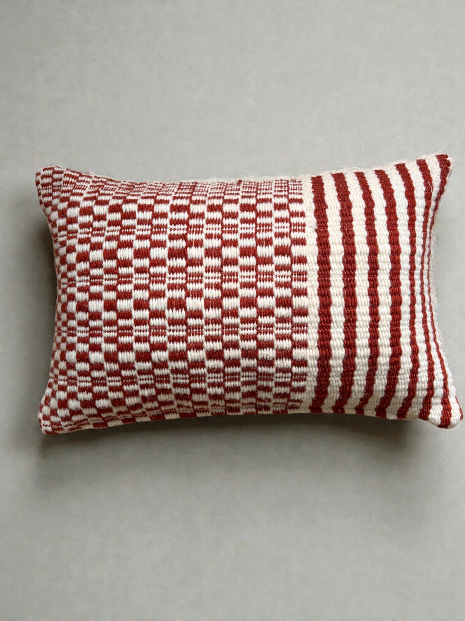 Das LEY! Heritage Line Kissen (40 x 60 cm) zeichnet sich durch einen gewebten rot-weißen Bezug mit geometrischen Mustern aus - links Schachbrettmuster, rechts Längsstreifen - und ist damit eine auffällige Wohnzimmerdeko auf neutralem Hintergrund.