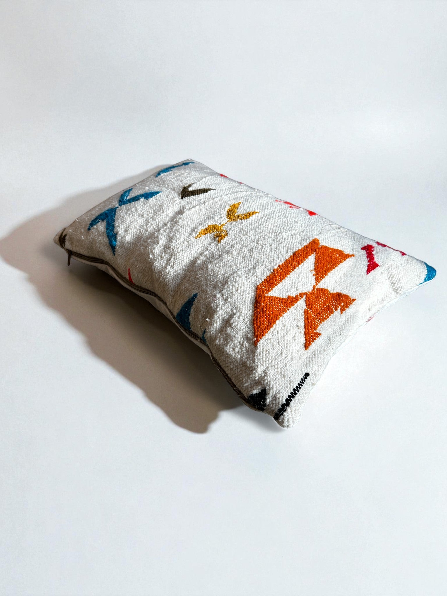 Das LEY! - Kissen - Colorful Line (40 x 60) von LEY! ist ein handgefertigtes, rechteckiges, weißes Kissen mit strukturiertem Stoff und lebendigen, abstrakten Formen in Orange, Gelb, Blau und Rot, die auf einer schlichten, weißen Oberfläche mit weichen Schatten dargestellt werden.