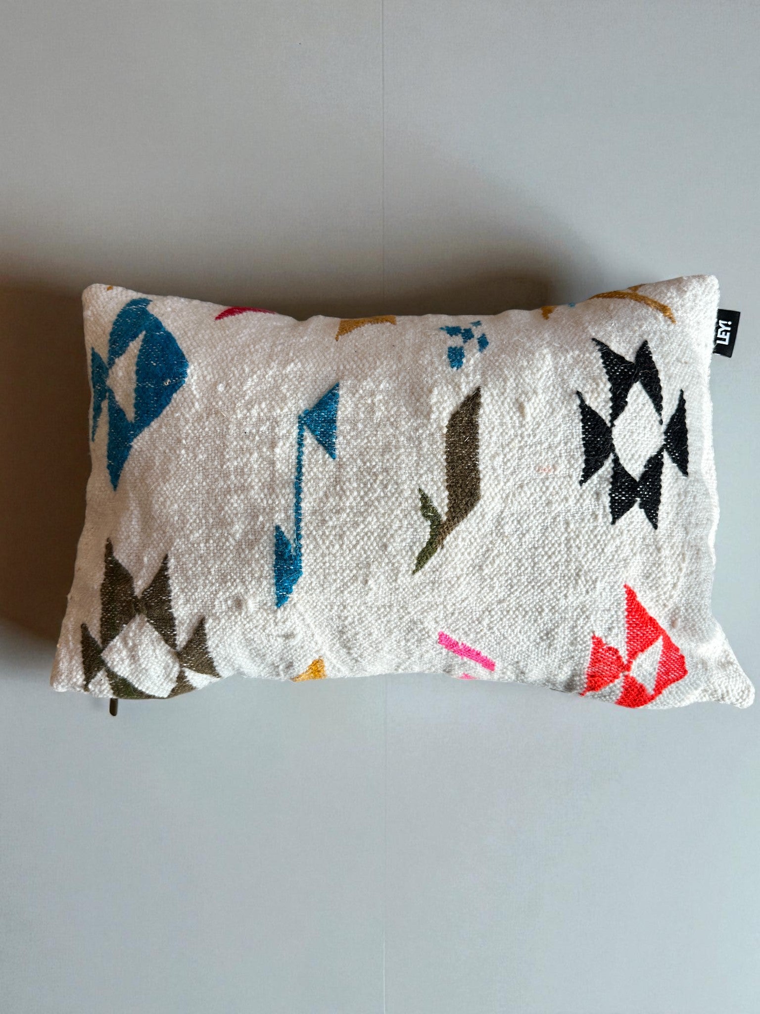 Das LEY! - Kissen - Colorful Line - 40 x 60 von LEY! ist ein rechteckiges, weißes, strukturiertes Kissen mit leuchtend blauen, grünen, schwarzen, orangen und roten geometrischen Mustern, das auf einer einfarbigen, hellgrauen Oberfläche liegt.