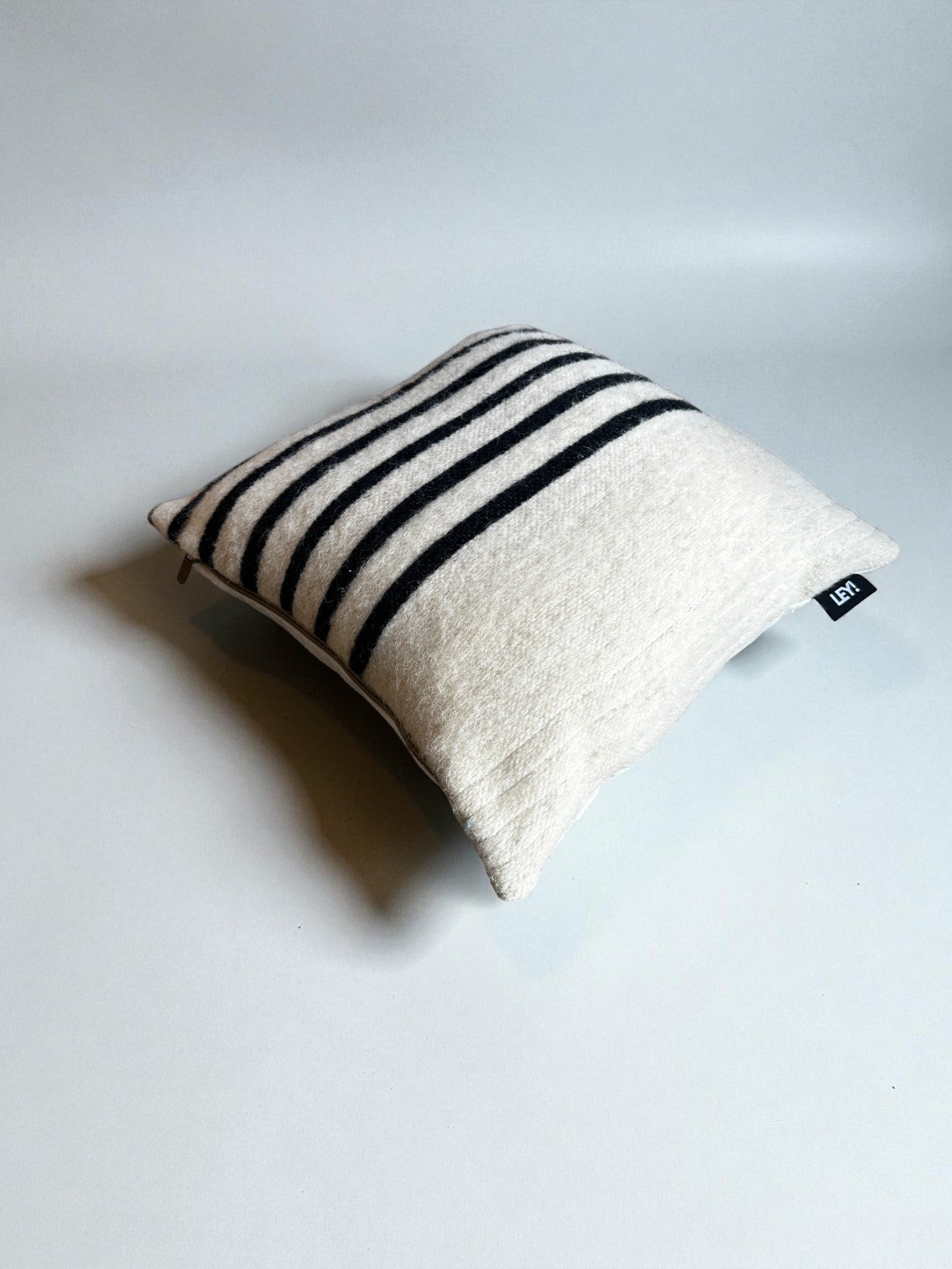 Ein quadratisches, cremefarbenes Kissen - WINTER EDITION - TAU - 40 x 40 von LEY! mit schwarzen Querstreifen und einem kleinen schwarzen Stoffanhänger liegt auf einer schlichten hellgrauen Fläche.