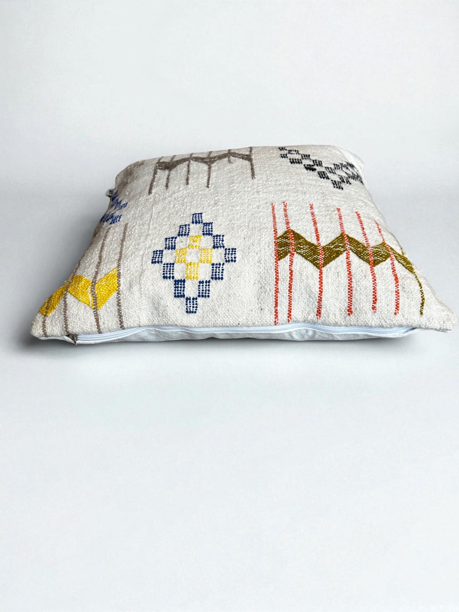 Das LEY! Kissen - Colorful Line (60 x 60) von LEY! ist ein quadratisches Dekokissen mit weißem Untergrund und leuchtend gelben, blauen, roten und grünen geometrischen Mustern, inspiriert vom handgemachten Marrakesch-Stil.