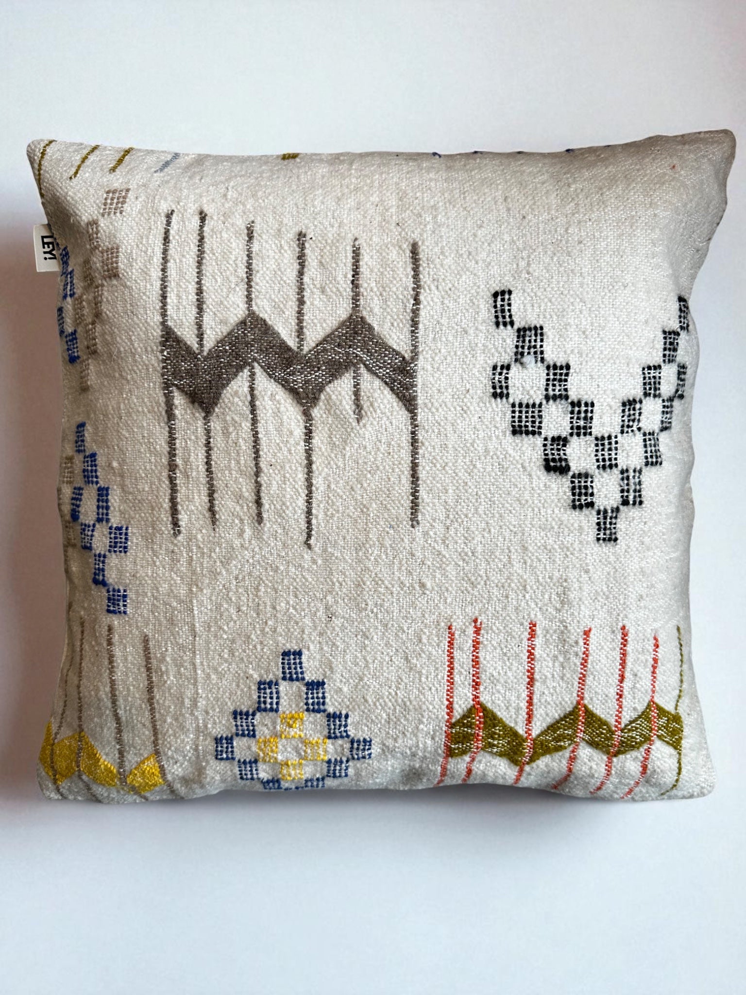 Das LEY! Kissen - Colorful Line (60x60 cm) der Marke LEY! ist ein quadratisches Kissen aus Schurwolle in Beige mit bunten geometrischen Mustern in Grau, Gelb, Rot und Blau sowie Zickzack-, Linien- und Rautendesign.