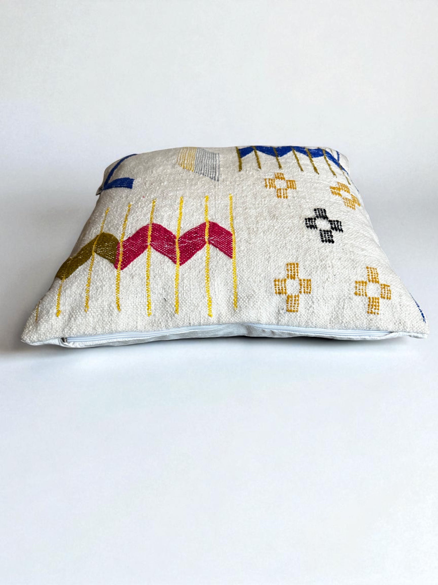 Das LEY! Kissen - Colorful Line - 60 x 60 von LEY! ist ein quadratisches Kissen aus weißer strukturierter Schurwolle, handgefertigt mit geometrischen Mustern in Gelb, Rosa, Blau und Schwarz, abgebildet auf einem einfarbig weißen Hintergrund.