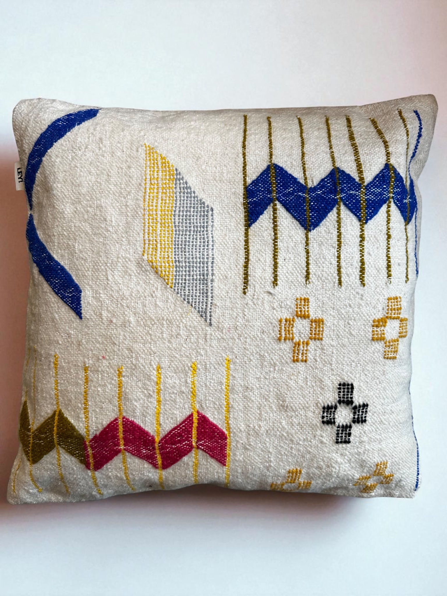 Das LEY! Kissen - Colorful Line (60x60) von LEY! ist ein quadratisches Kissen aus strukturiertem weißem Schurwolle-Stoff mit handgestickten blauen, gelben, rosa, grauen und schwarzen geometrischen Mustern - Zickzack, Streifen und Zeichen - auf einem unifarbenen Hintergrund.